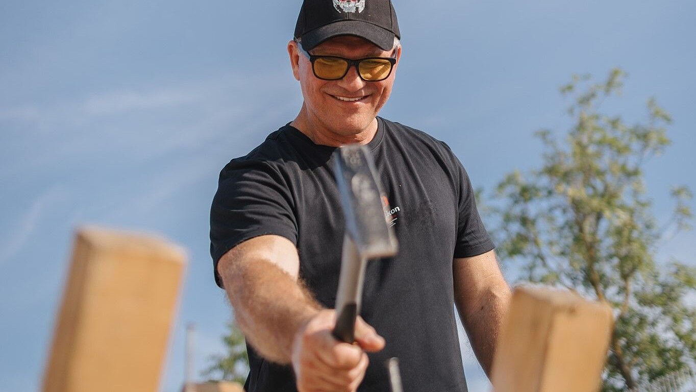 Man wearing sunglasses and a baseball cap, working with a hammer on wood, in front of a blue sky and trees.