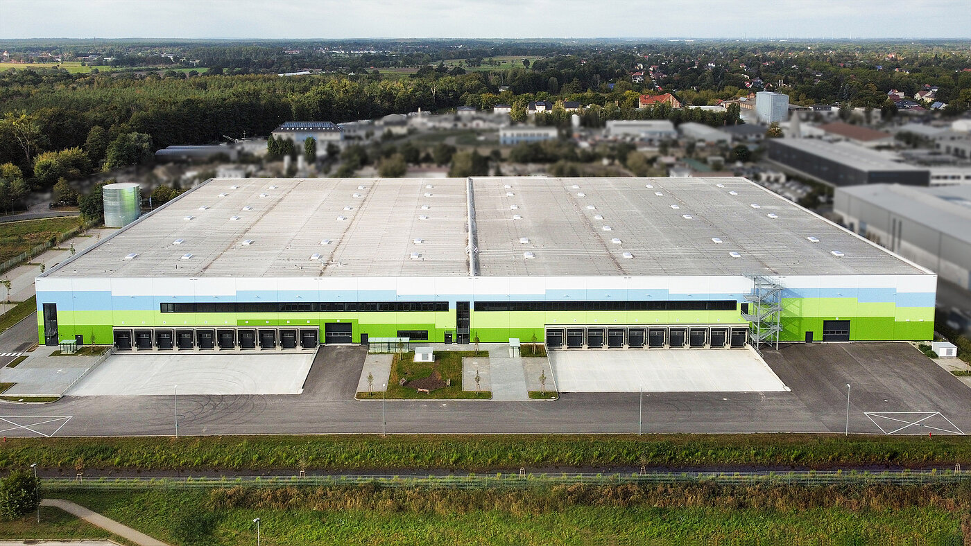 Große Lagerhalle mit grüner und blauer Fassade, Lastabfertigungsbereich und angrenzender Landschaft.