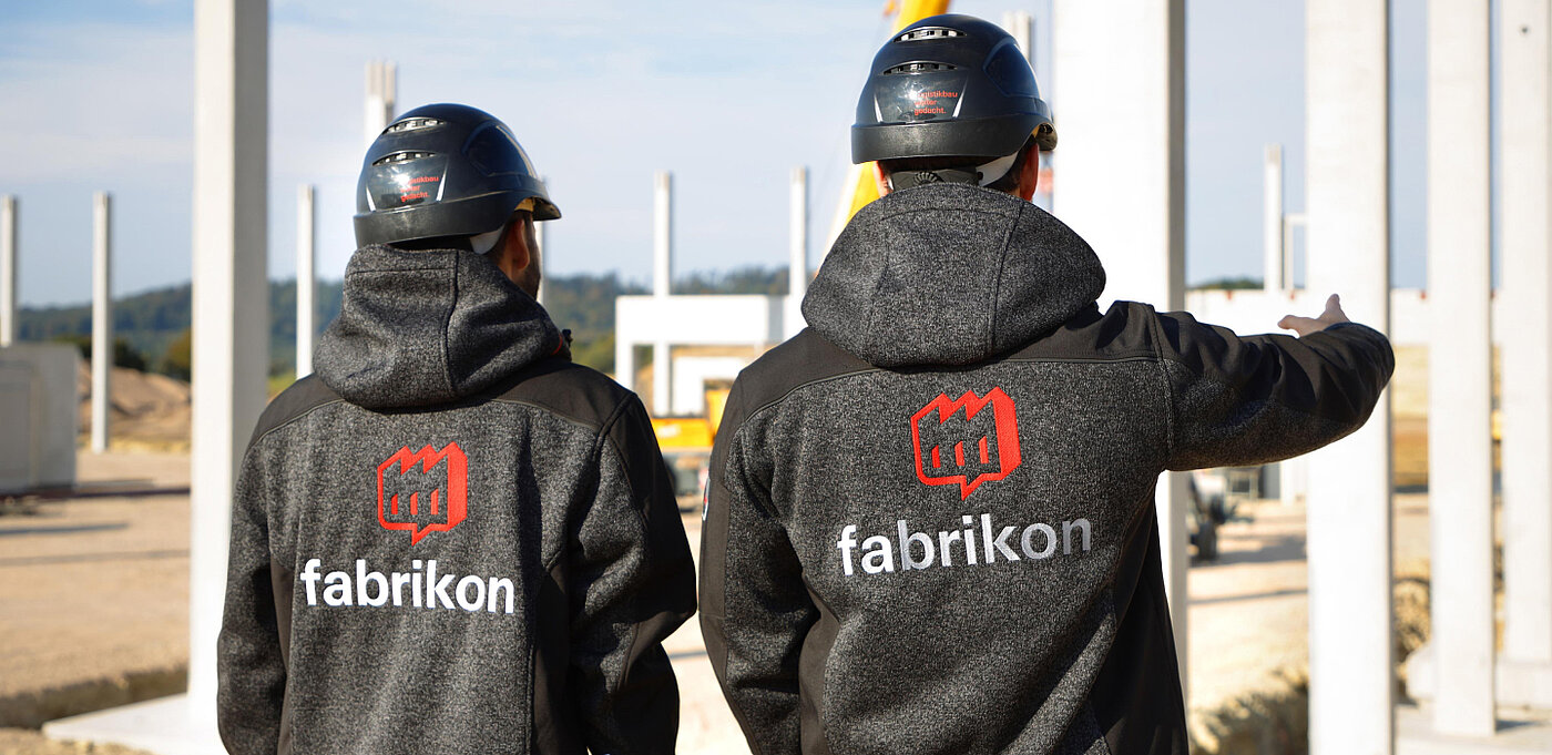 Zwei Personen in Helmen und Arbeitskleidung stehen auf einer Baustelle und zeigen auf die Baustelle.