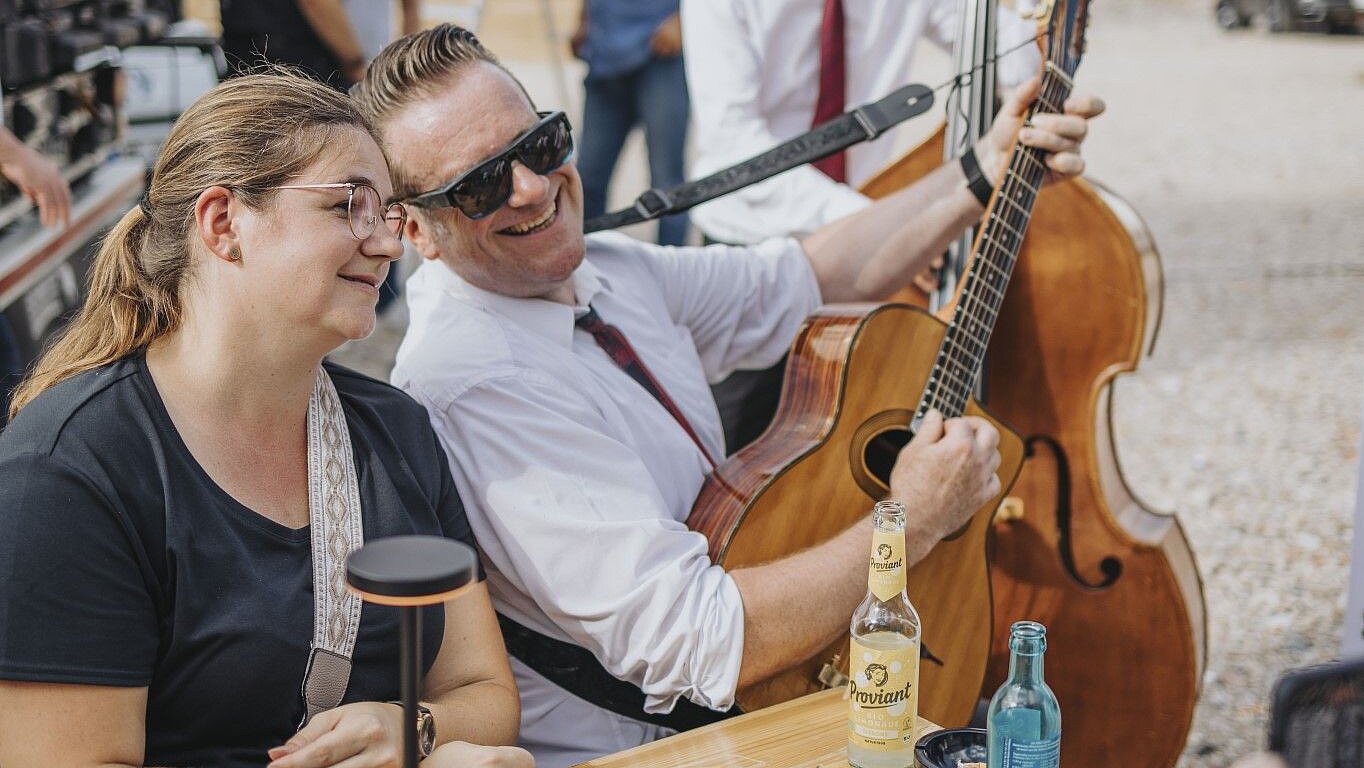A man with a guitar smiles next to a woman at a table, surrounded by drinks and a relaxed atmosphere.