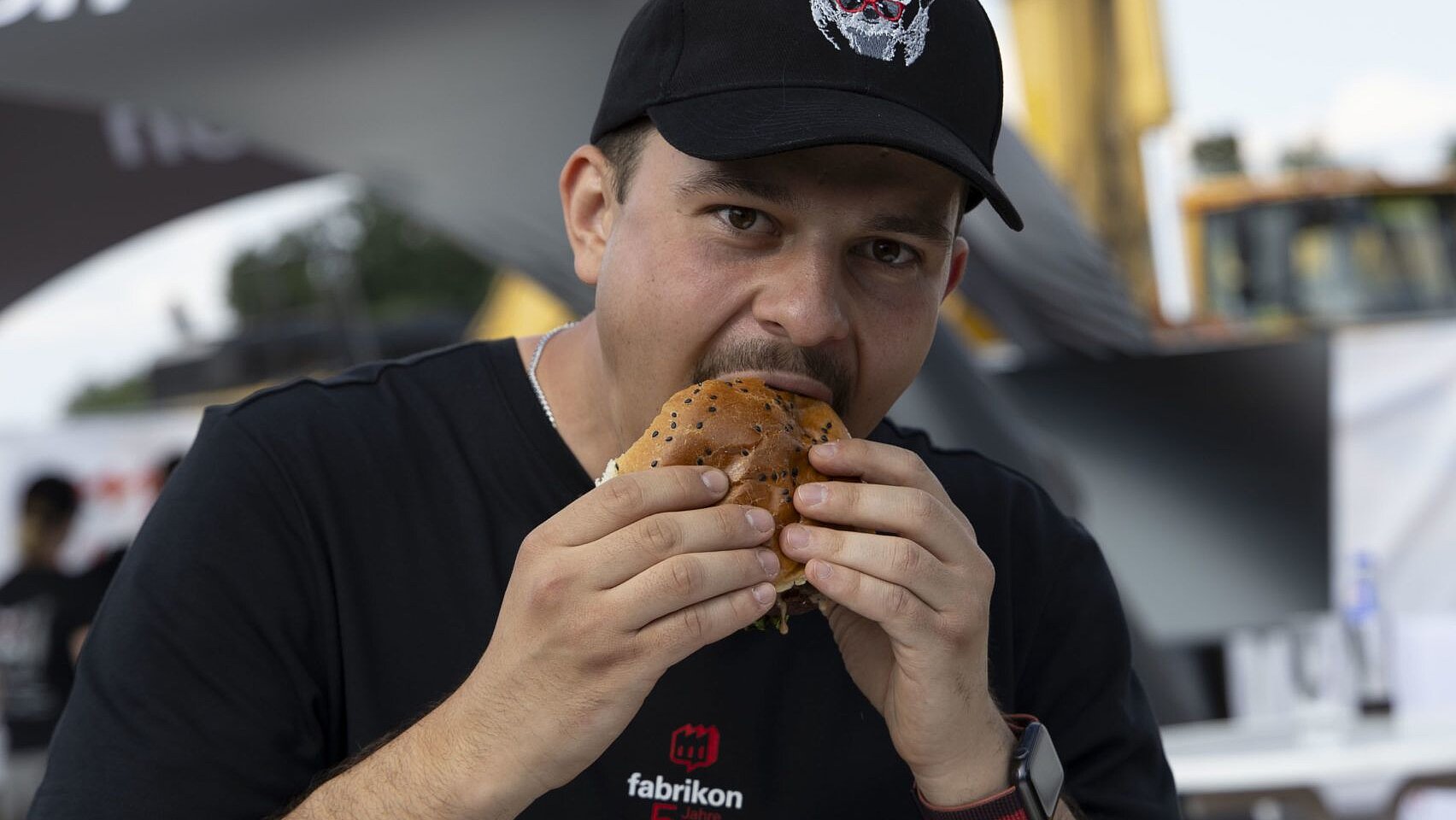 Man wearing a black T-shirt and cap is holding a hamburger and preparing to eat it.