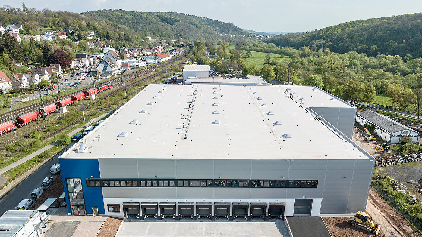 Luftaufnahme eines modernen Lagergebäudes mit mehreren Lkw-Rampen, umgeben von Natur und Wohnhäusern.