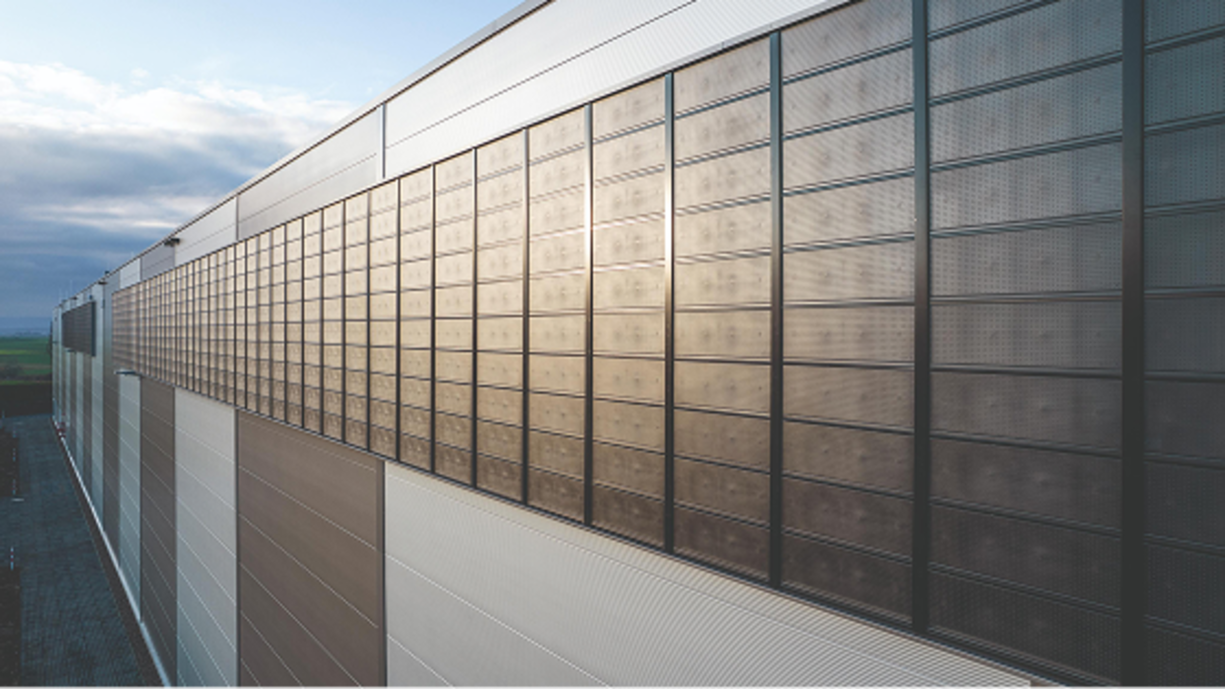 Fassade eines modernen Gebäudes mit horizontalen Elementen und großen Fenstern unter einem bewölkten Himmel.