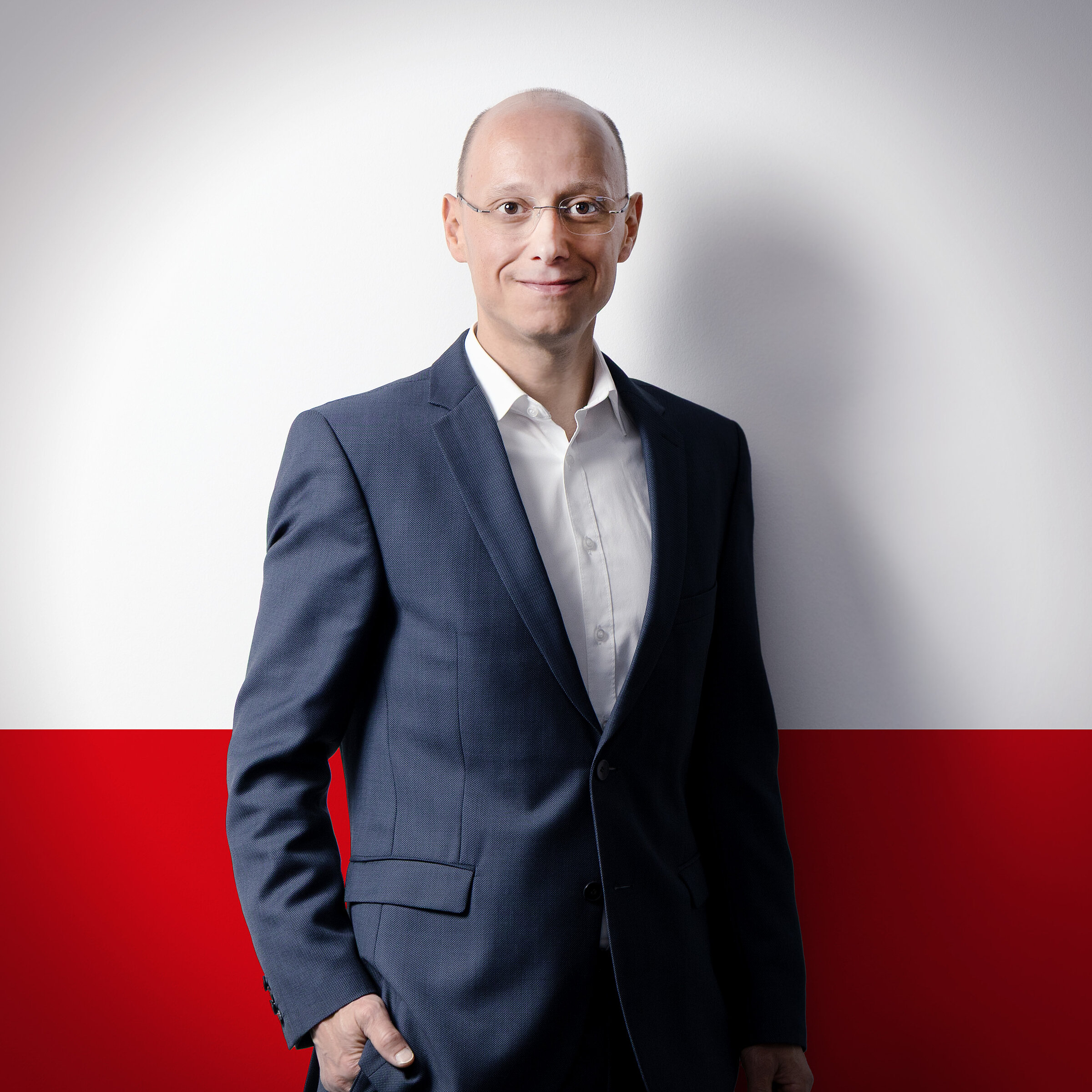 A man in a suit stands in front of a half-white, half-red wall and smiles at the camera.