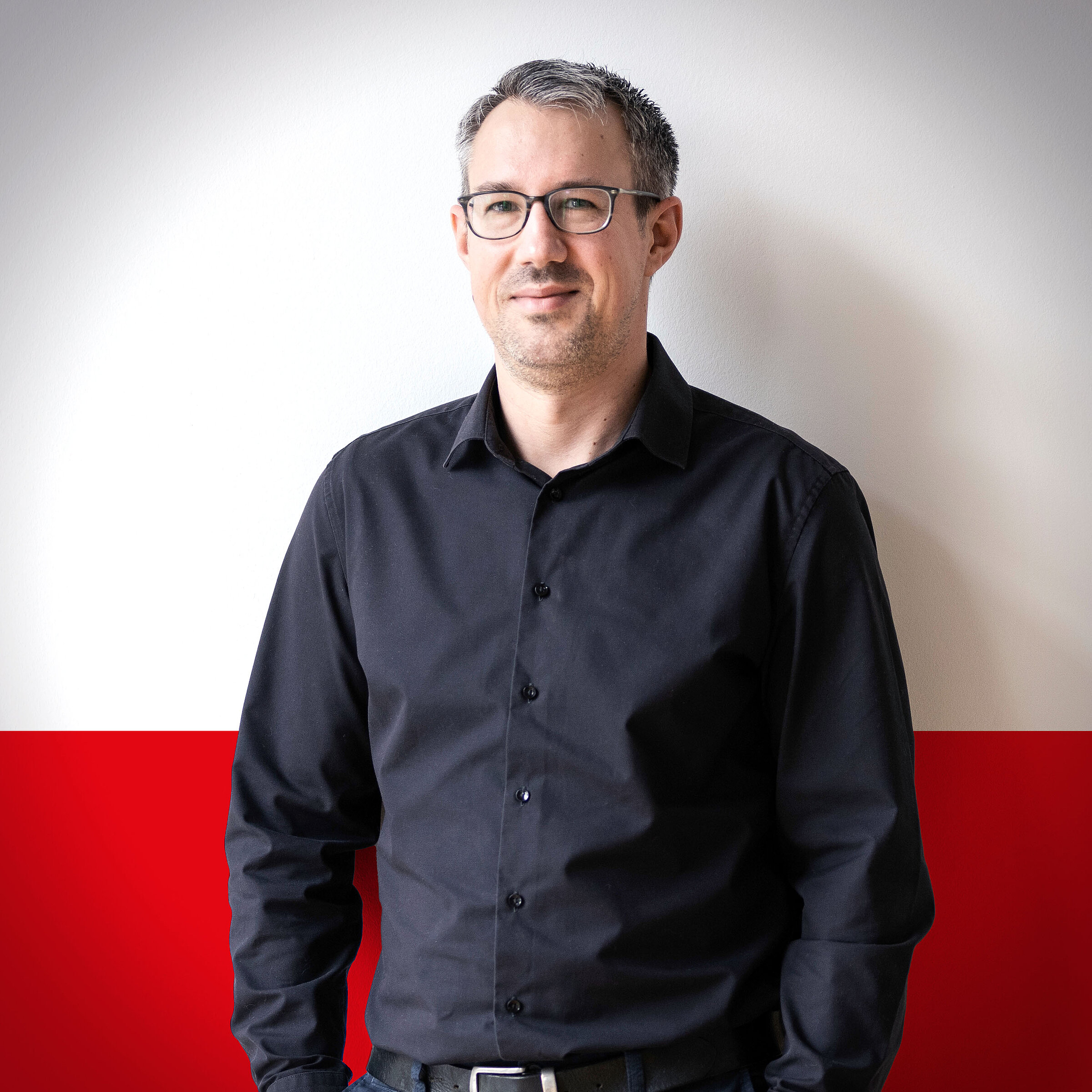Person in a black shirt with glasses standing in front of a white and red background.