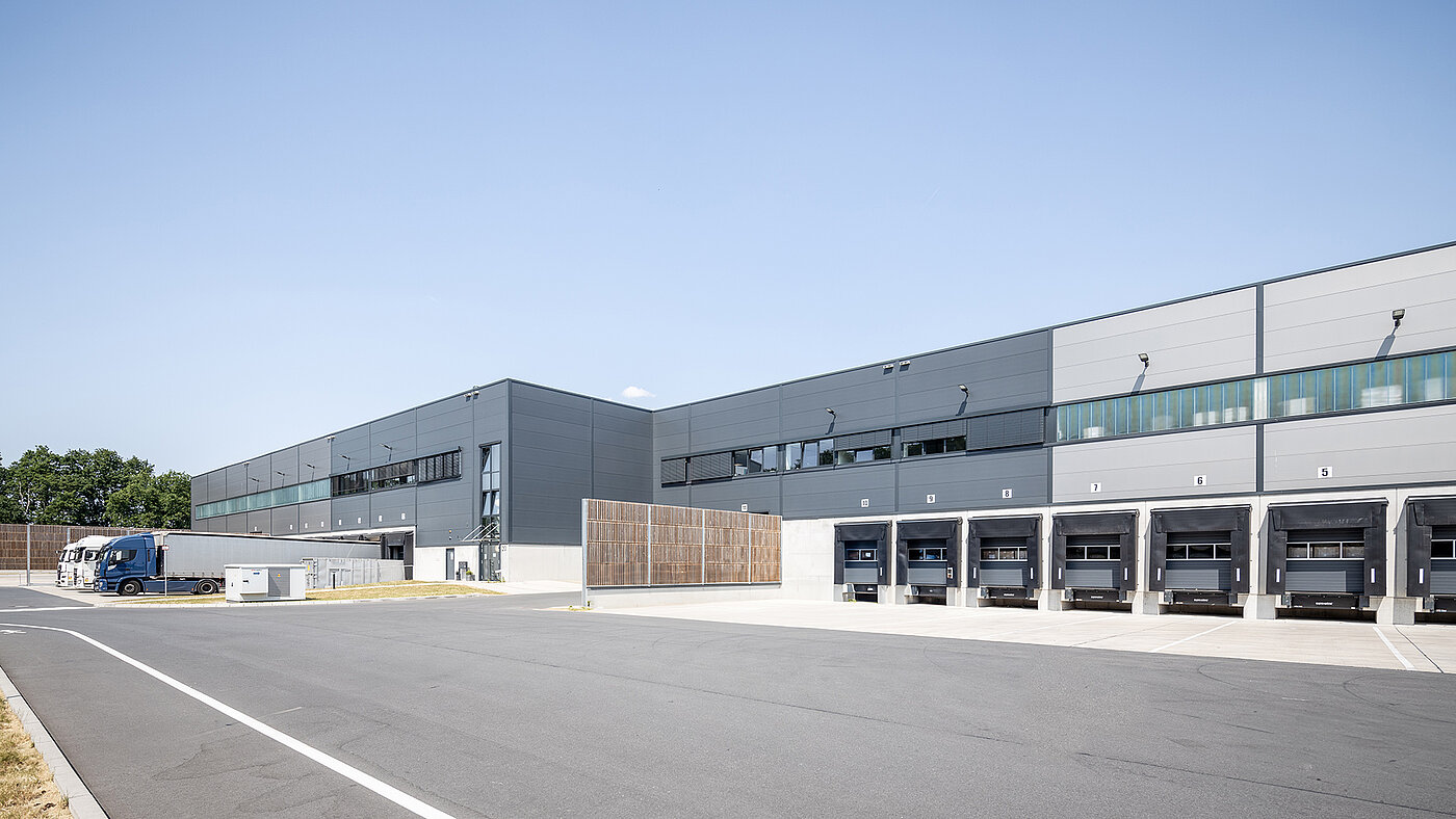 Moderne Lagerhalle mit Lkw-Entladezonen und großen Fenstern unter blauem Himmel.