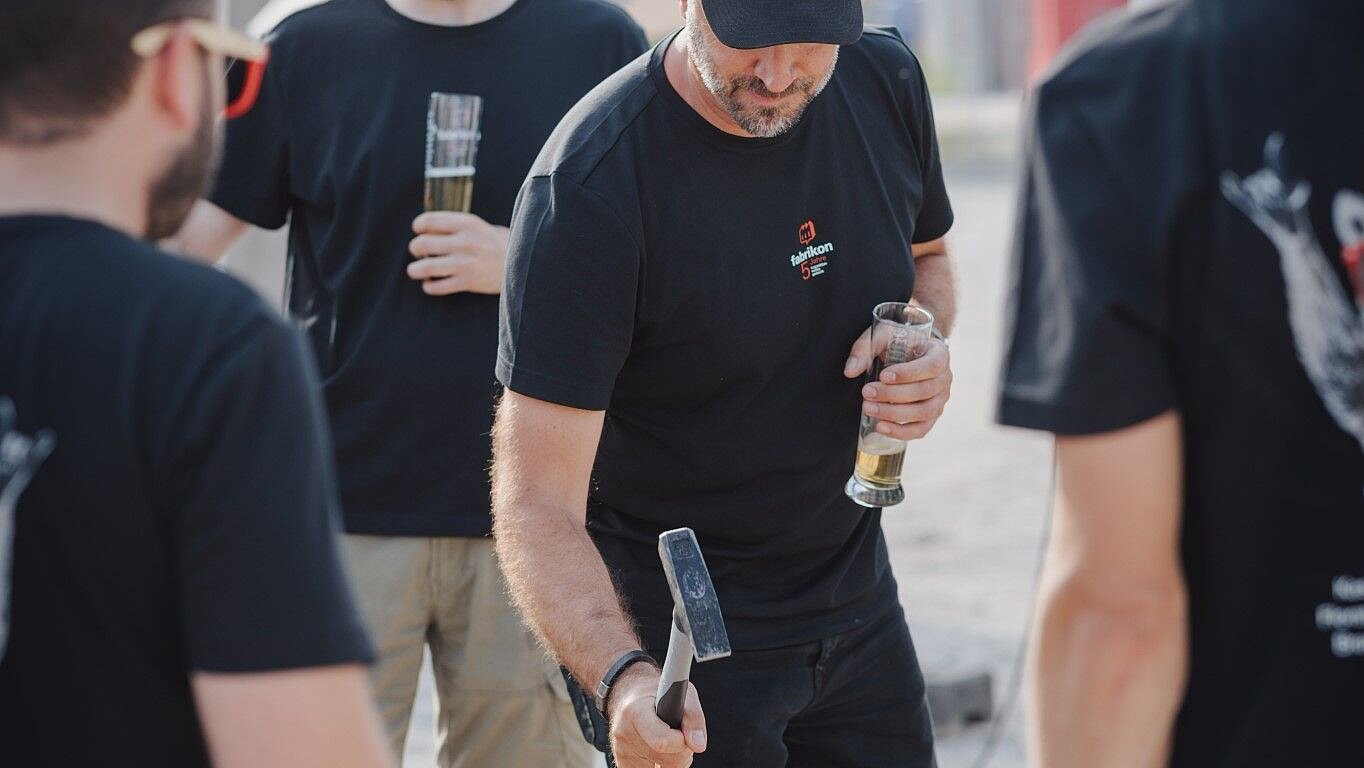 Men in T-shirts are working with hammers on a wooden structure to drive in nails. Background with tents and other people.