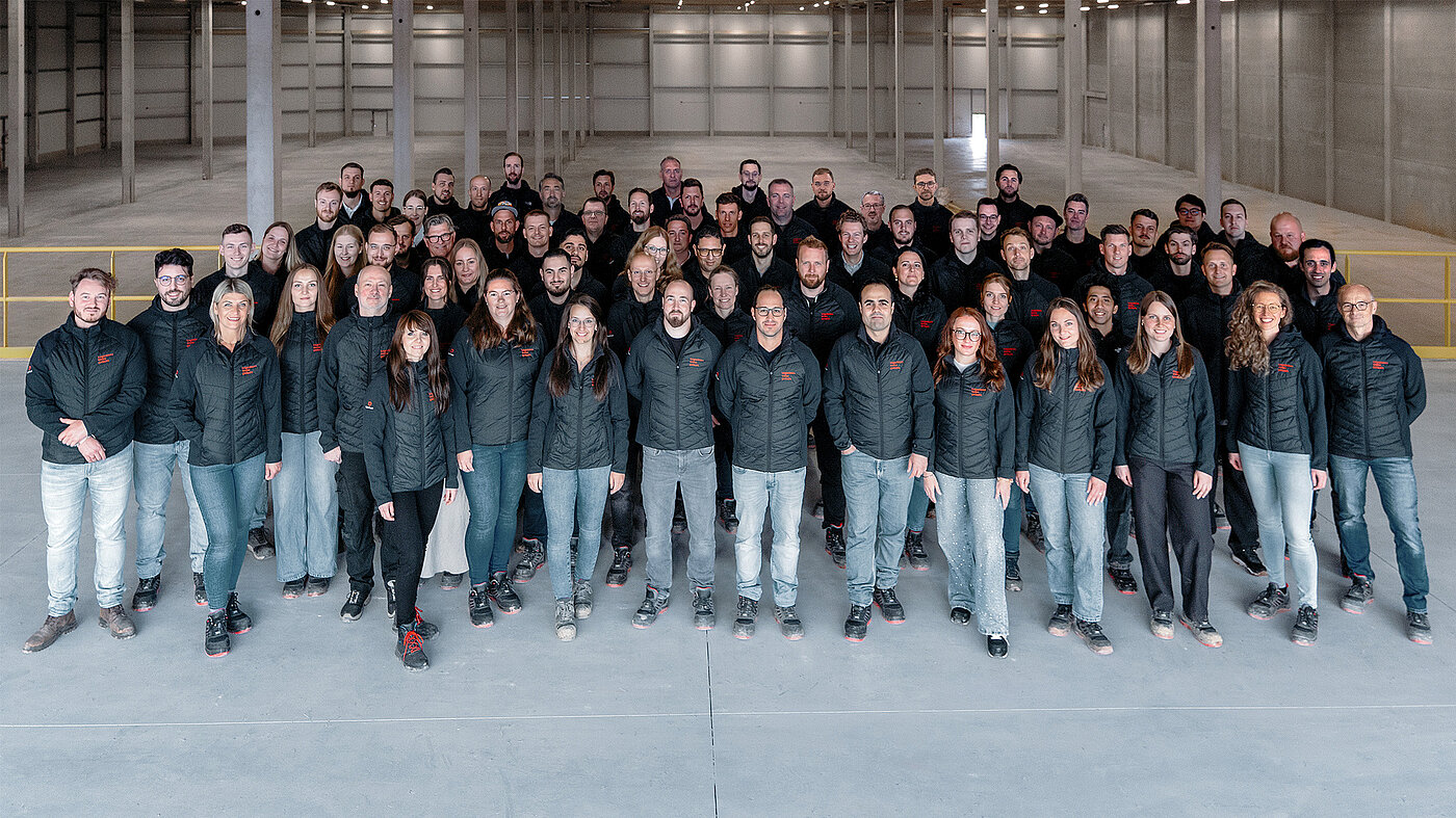 Gruppenfoto von fabrikon Mitarbeitenden, die auf einer Mezzanine in einer leeren Logistikhalle stehen und freundlich in die Kamera schauen.