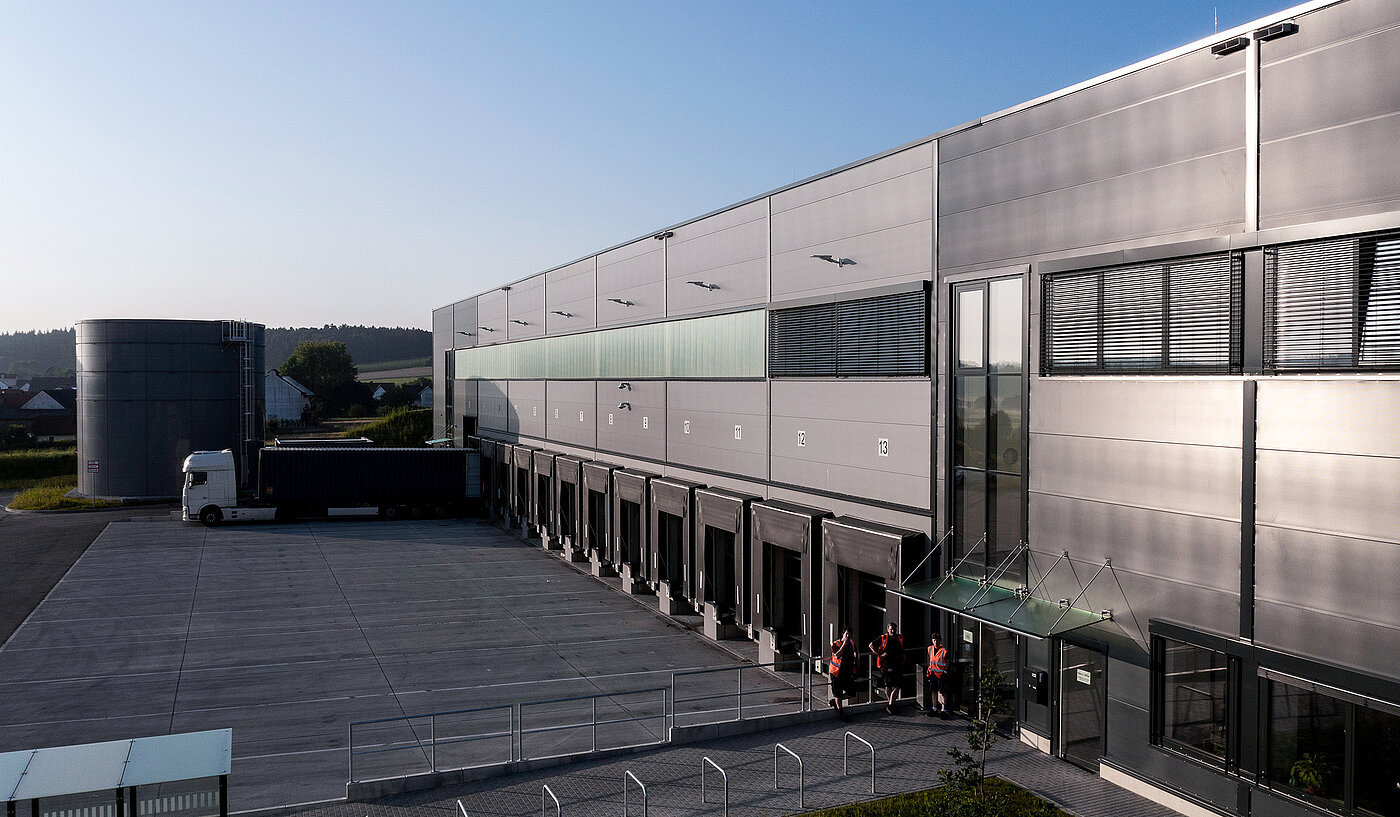 Moderne Lagerhalle mit Ladebereichen, einem Lkw und Angestellten in Warnwesten an der Eingangstür.