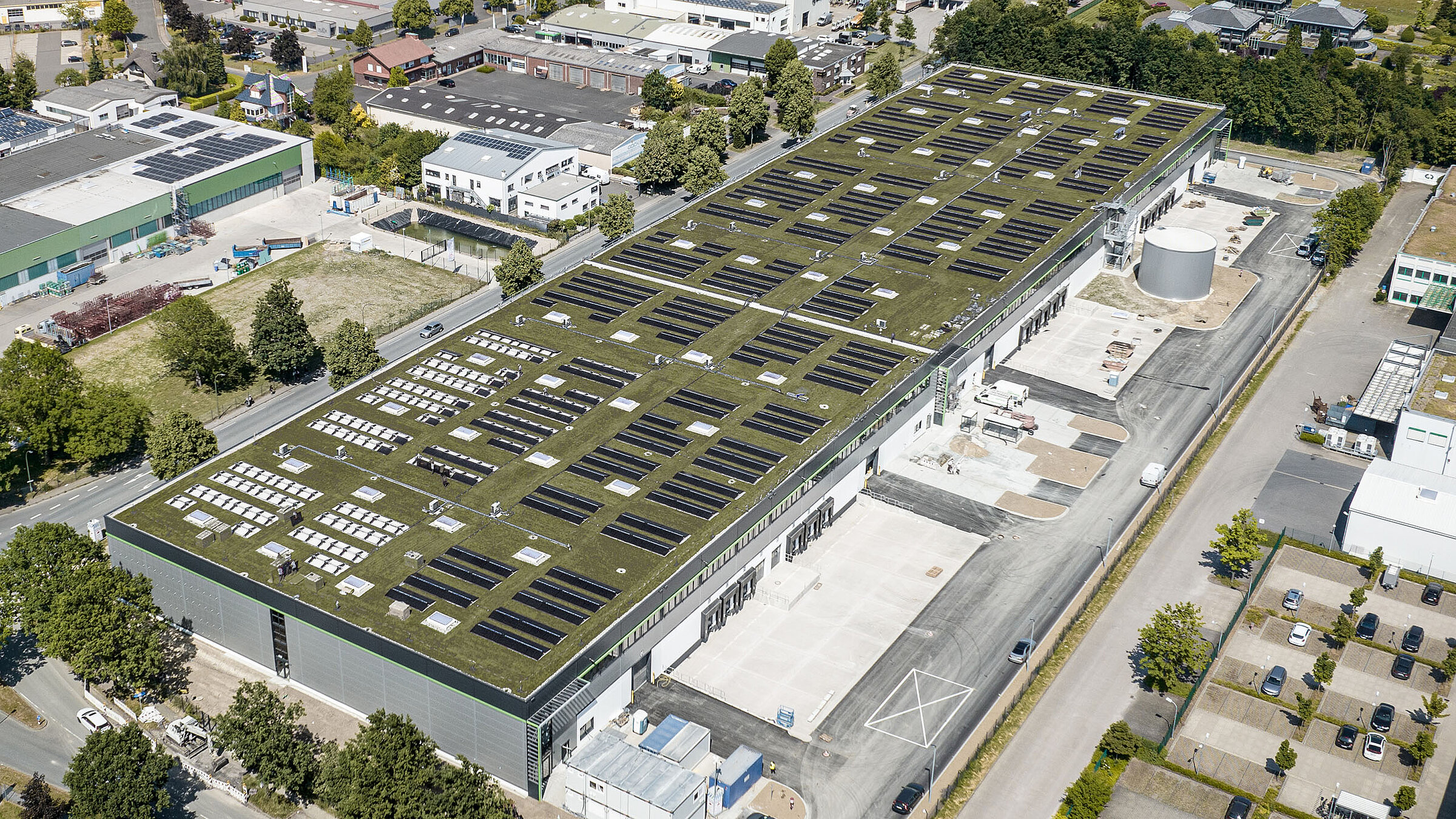 Luftaufnahme eines modernen Industriegebäudes mit grüner Dachbegrünung und Solarpanelen. Umgebung mit Bäumen und Straßen.