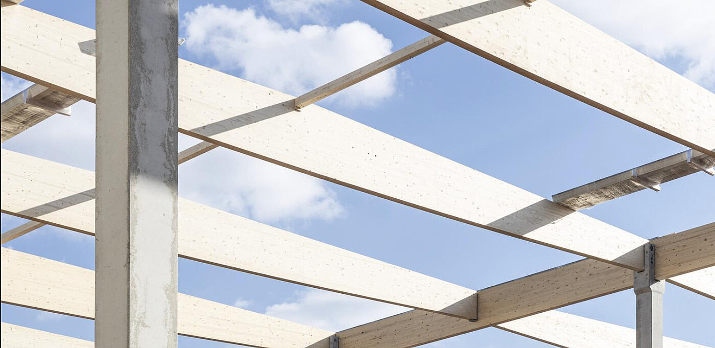 Blick auf eine moderne Gebäudestruktur mit Holzlamellen und Betonstützen vor einem blauen Himmel mit Wolken.