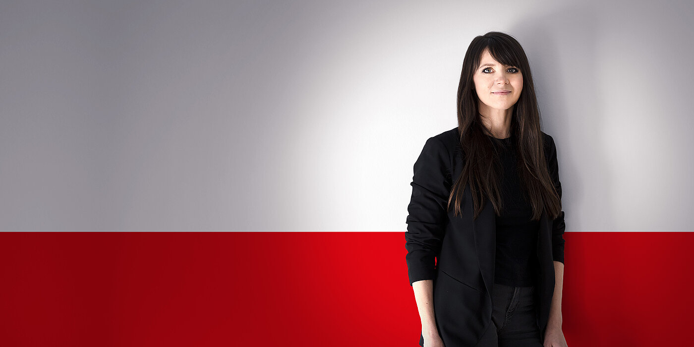 Porträt einer Frau in schwarzer Kleidung vor einem grau-roten Hintergrund. Sie hat lange, dunkle Haare und blickt in die Kamera.