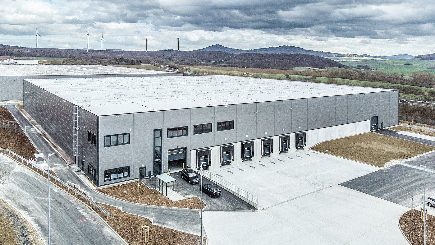 Großes modernes Lagergebäude mit Ladebucht, umgeben von Landschaft und Windkraftanlagen im Hintergrund.