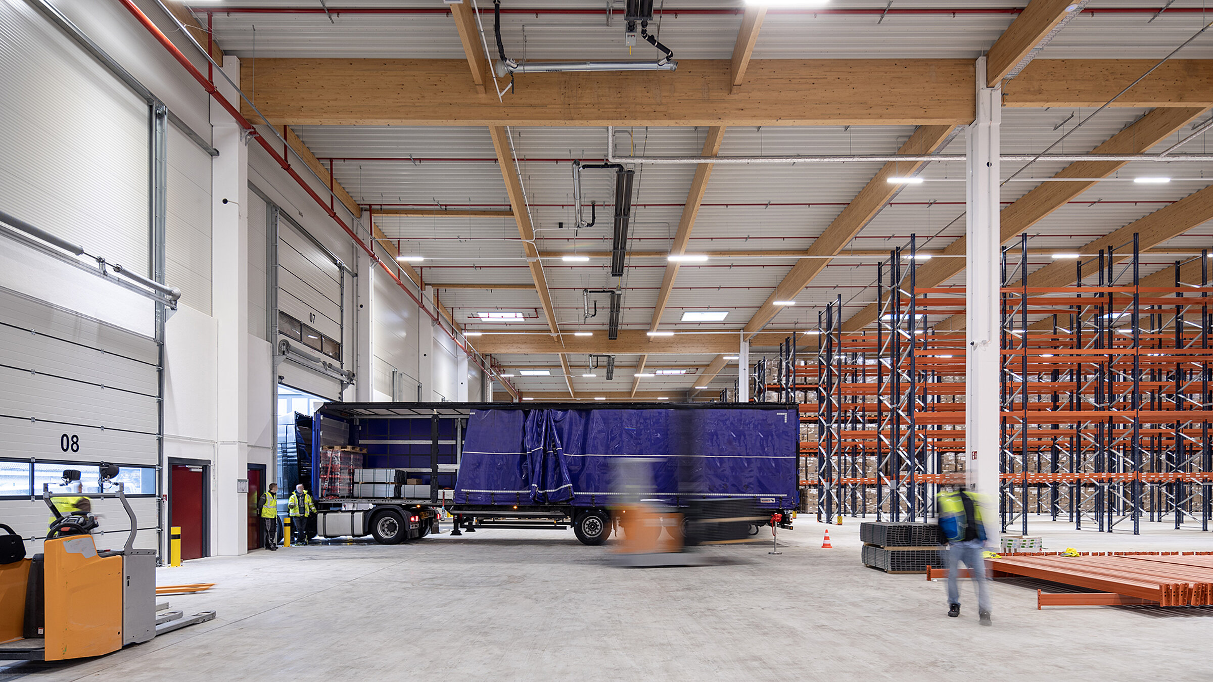 Lagerhalle mit Lkw an der Rampe, Mitarbeiter in Sicherheitskleidung, Regale im Hintergrund, Arbeitsumgebung.
