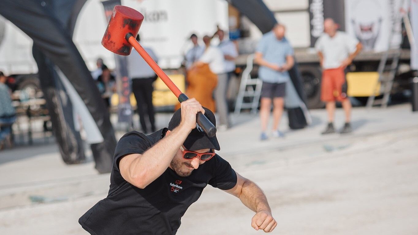 A man swings a heavy hammer above his head and prepares to strike a block on the ground.