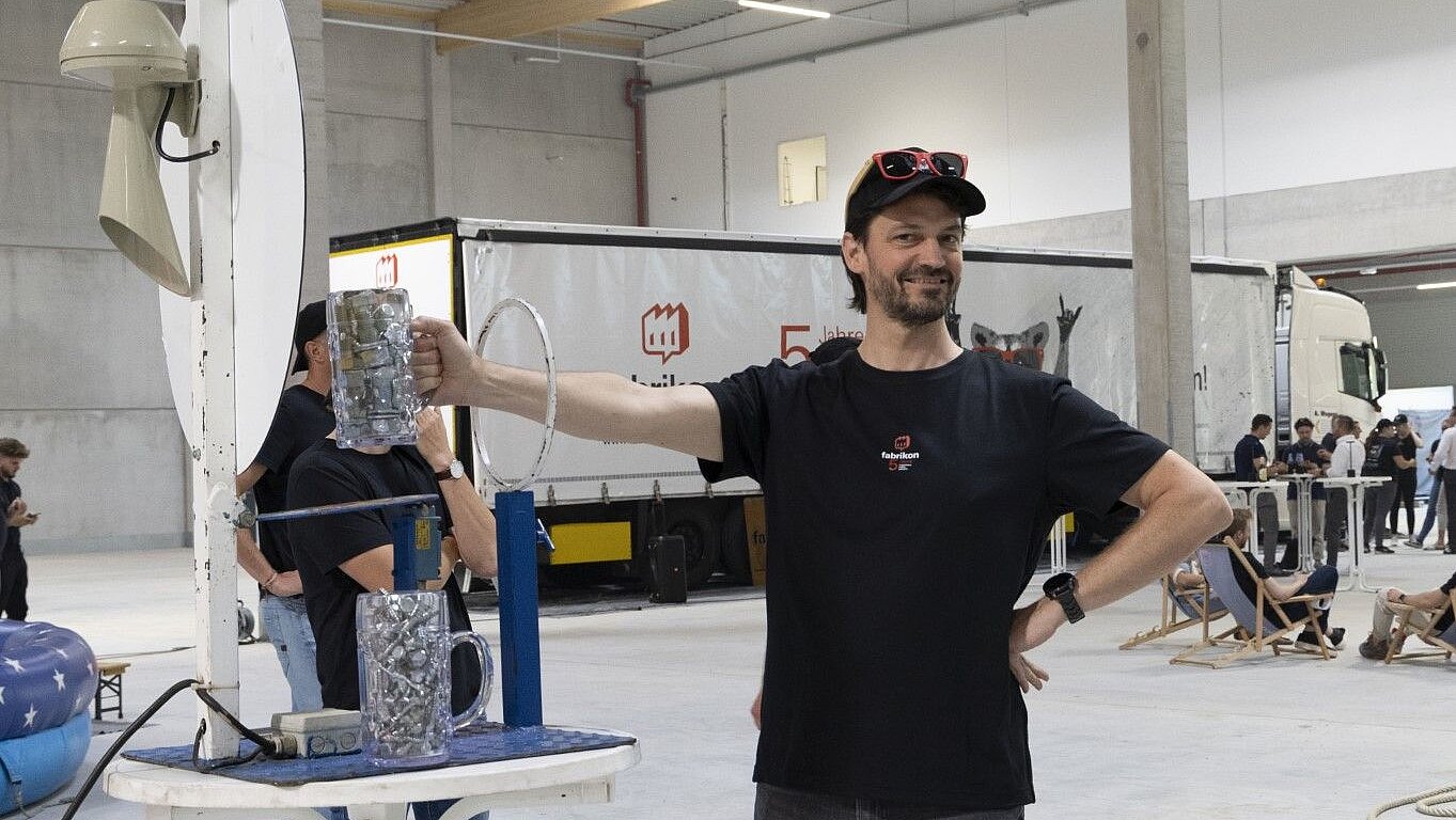 Man wearing a black T-shirt and sunglasses holds a container in a warehouse-like setting, next to a table game.