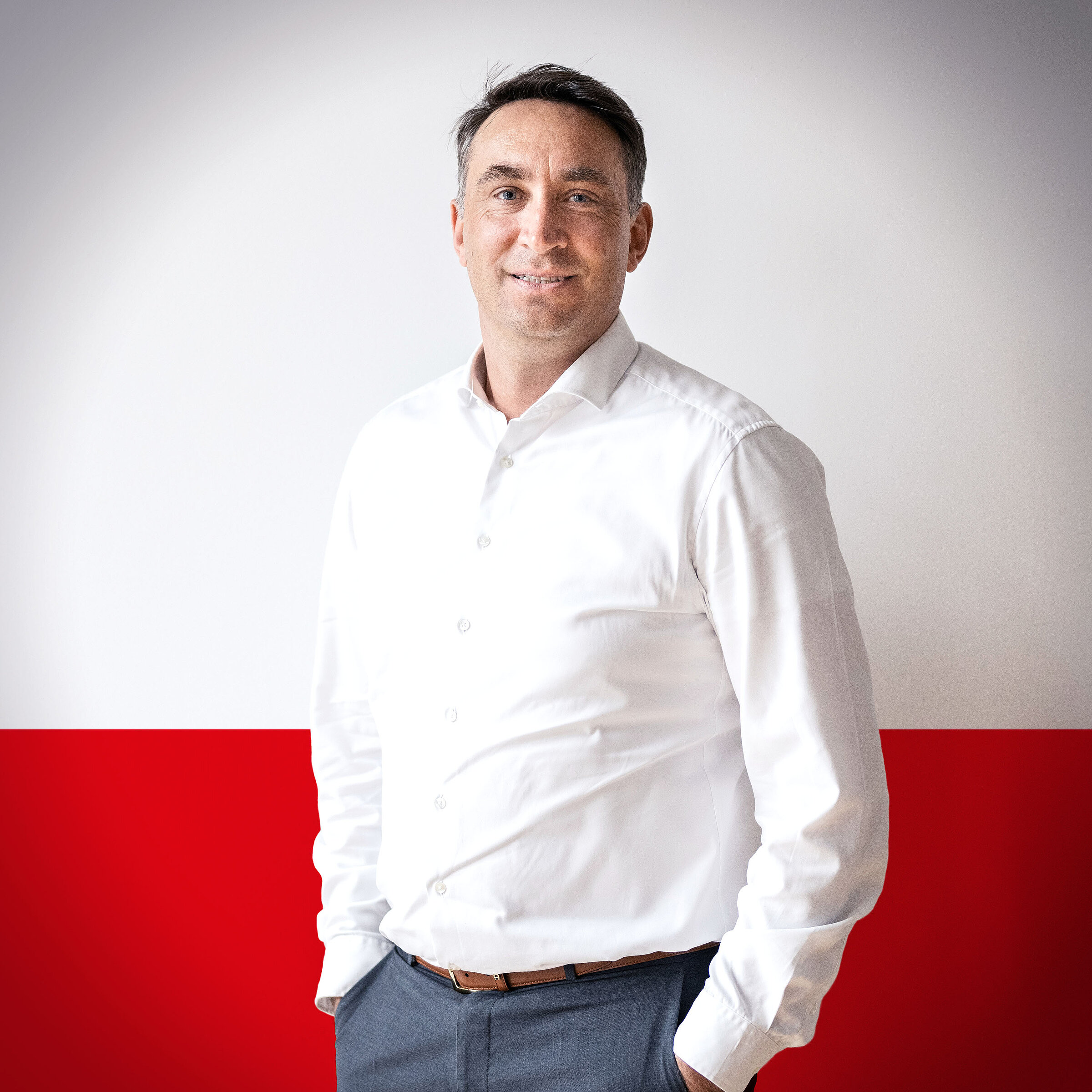 Portrait of a smiling man in a white shirt with a red background. He is standing relaxed with both hands in his pockets.