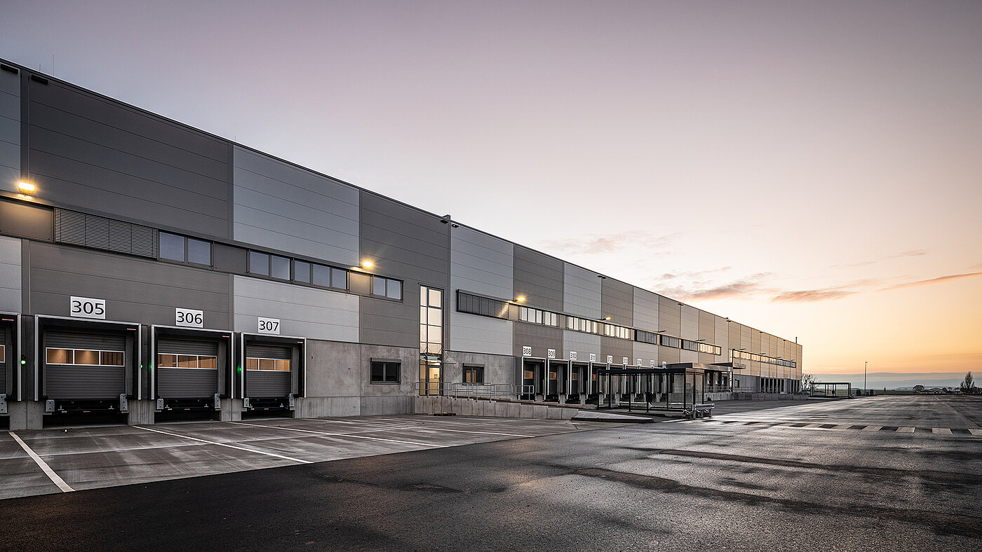 Moderne Lagerhalle bei Sonnenaufgang mit Laderampen und Parkplätzen im Vordergrund.