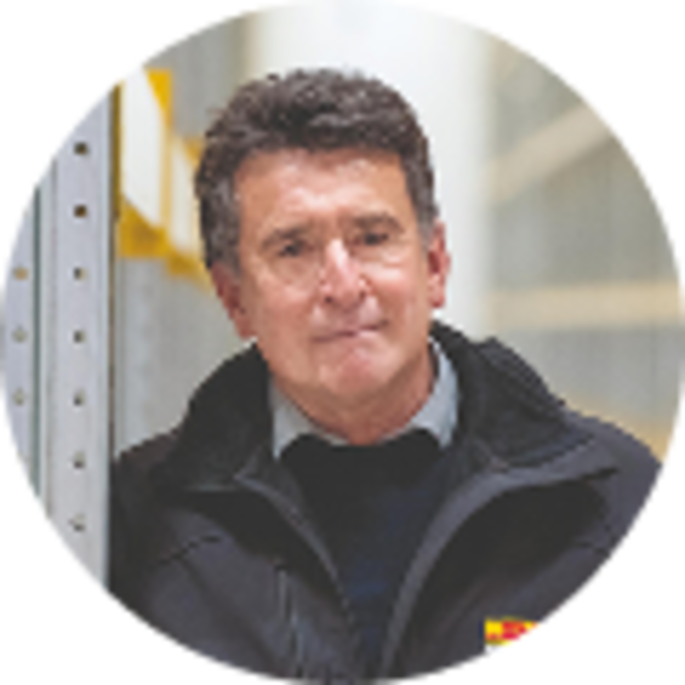 Mann mit kurzen, dunklen Haaren und einer schwarzen Jacke, lächelt, steht vor einer hellen, industriellen Hintergrundumgebung.