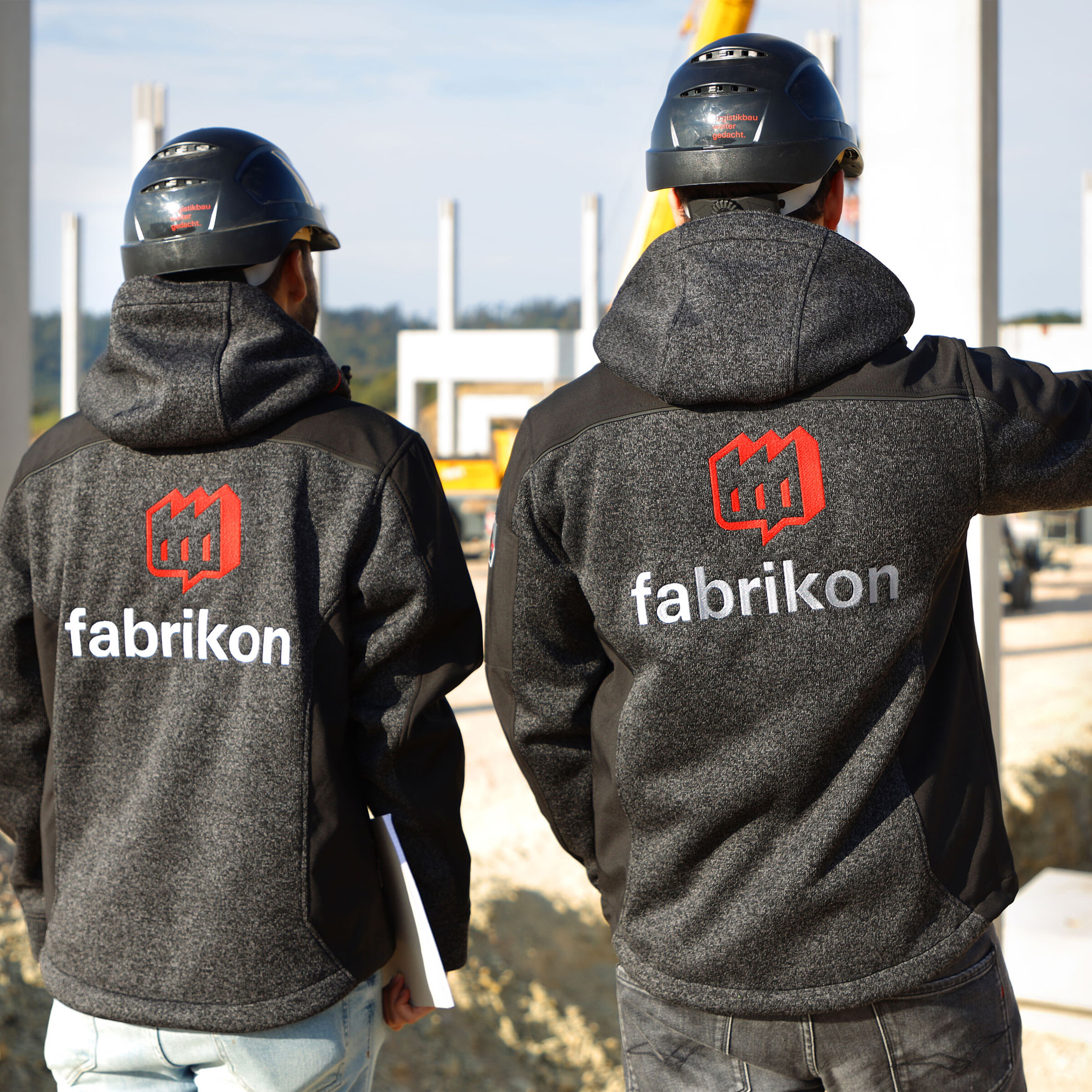 Zwei Personen mit Schutzhelmen stehen auf einer Baustelle und blicken in Richtung der Rohbauten.