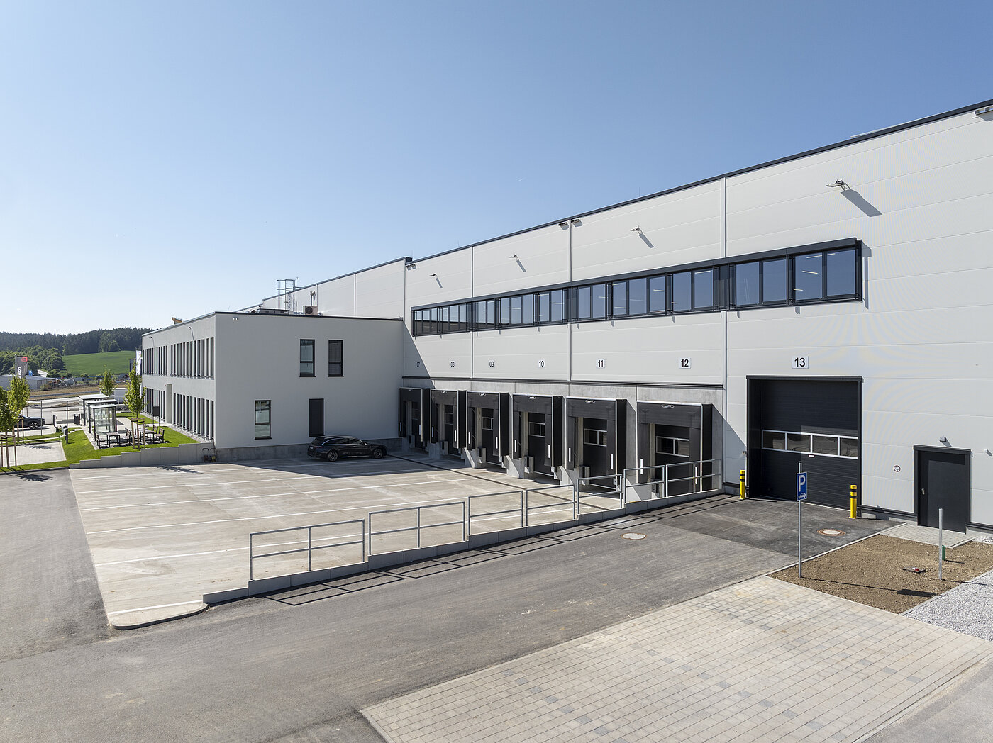 Modernes Lagergebäude mit Ladebuchten, Parkplatz und Grünflächen, bei klarem blauen Himmel.