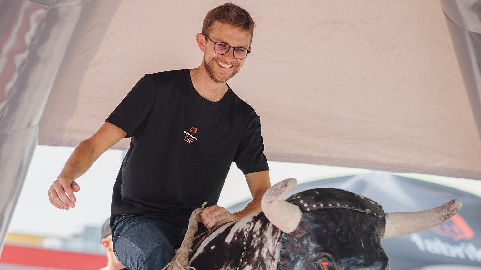 Man wearing glasses riding a mechanical bull under a tent. Fun and action at an event.