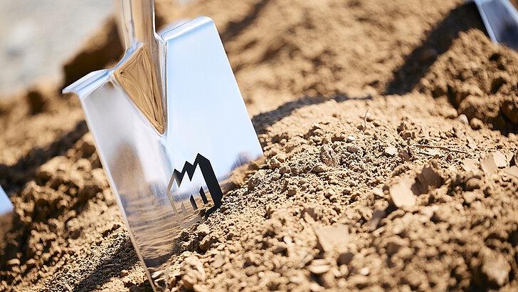 A shiny spade stands on dry earth, ready for digging, with a logo on the blade.
