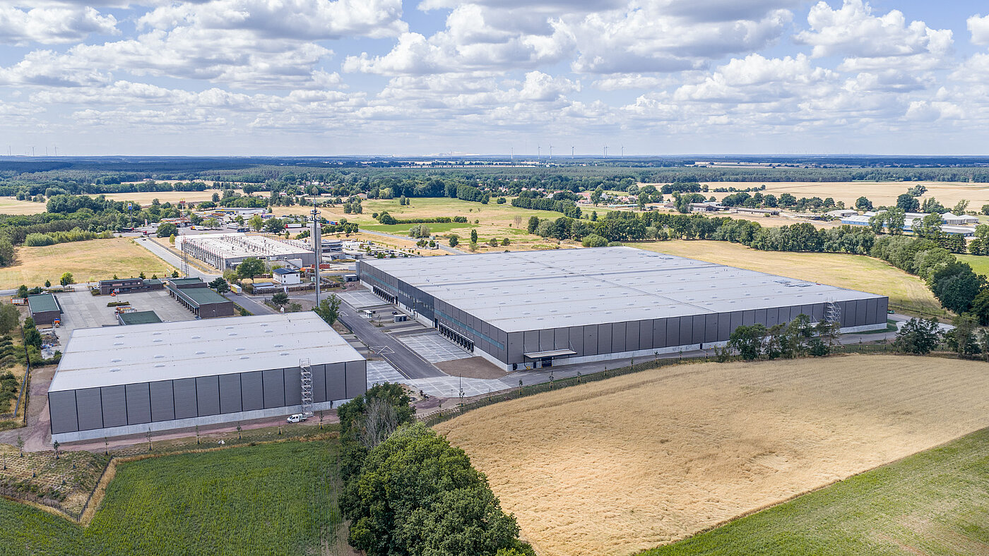 Luftaufnahme eines modernen Lagerhauses mit grünen Feldern und Wolken am Himmel. Vielfältige Landschaft im Hintergrund.