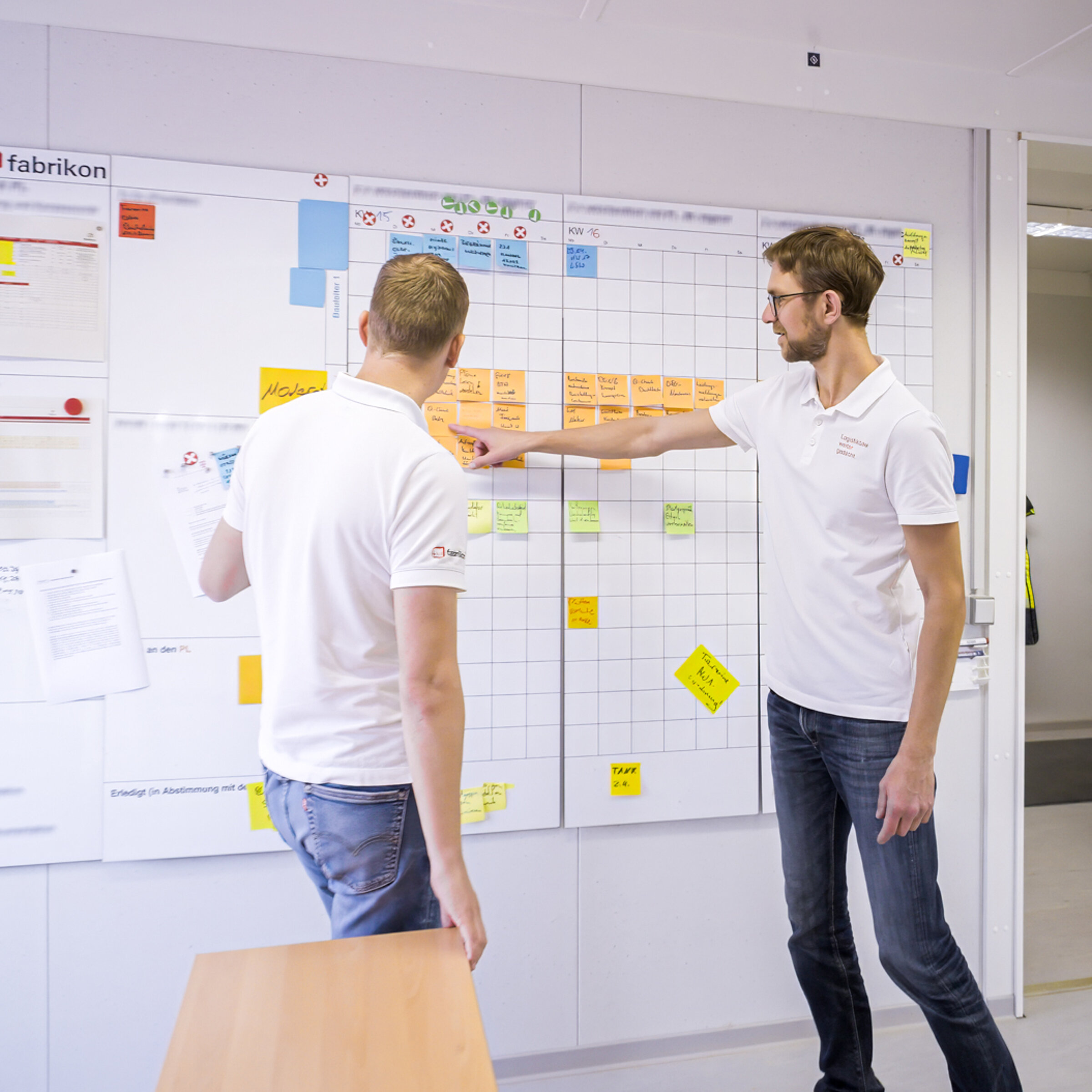 Zwei Männer mit Hemden diskutieren vor einem großen Planungsboard mit bunten Notizen und Grafiken in einem Büro.