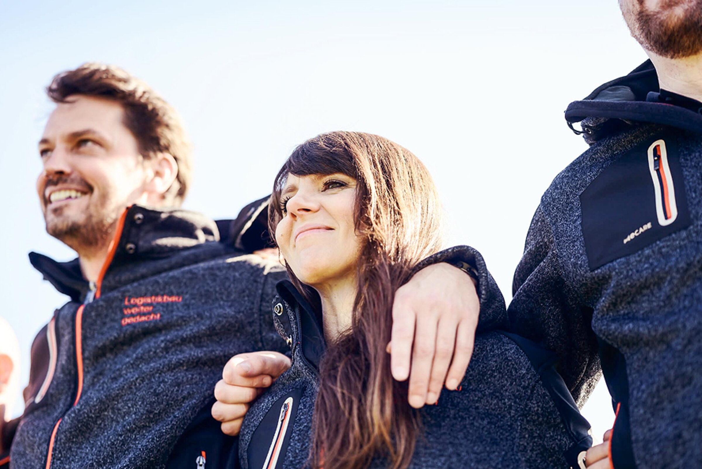 Drei Personen stehen zusammen, lächelnd in warmen Jacken, unter einem blauen Himmel. Freundliche Atmosphäre.
