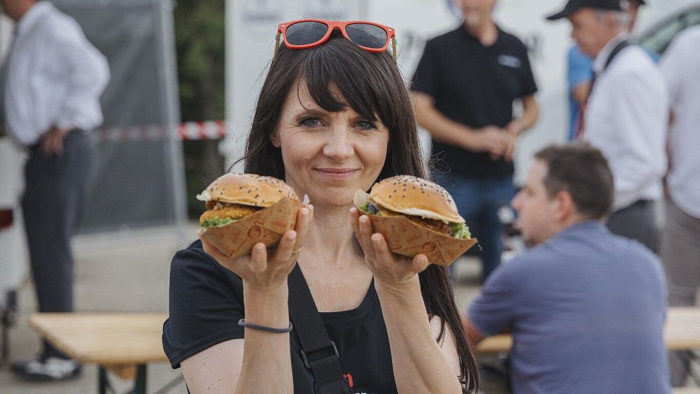 A person is holding two hamburgers in their hands, with other people and food stalls in the background.