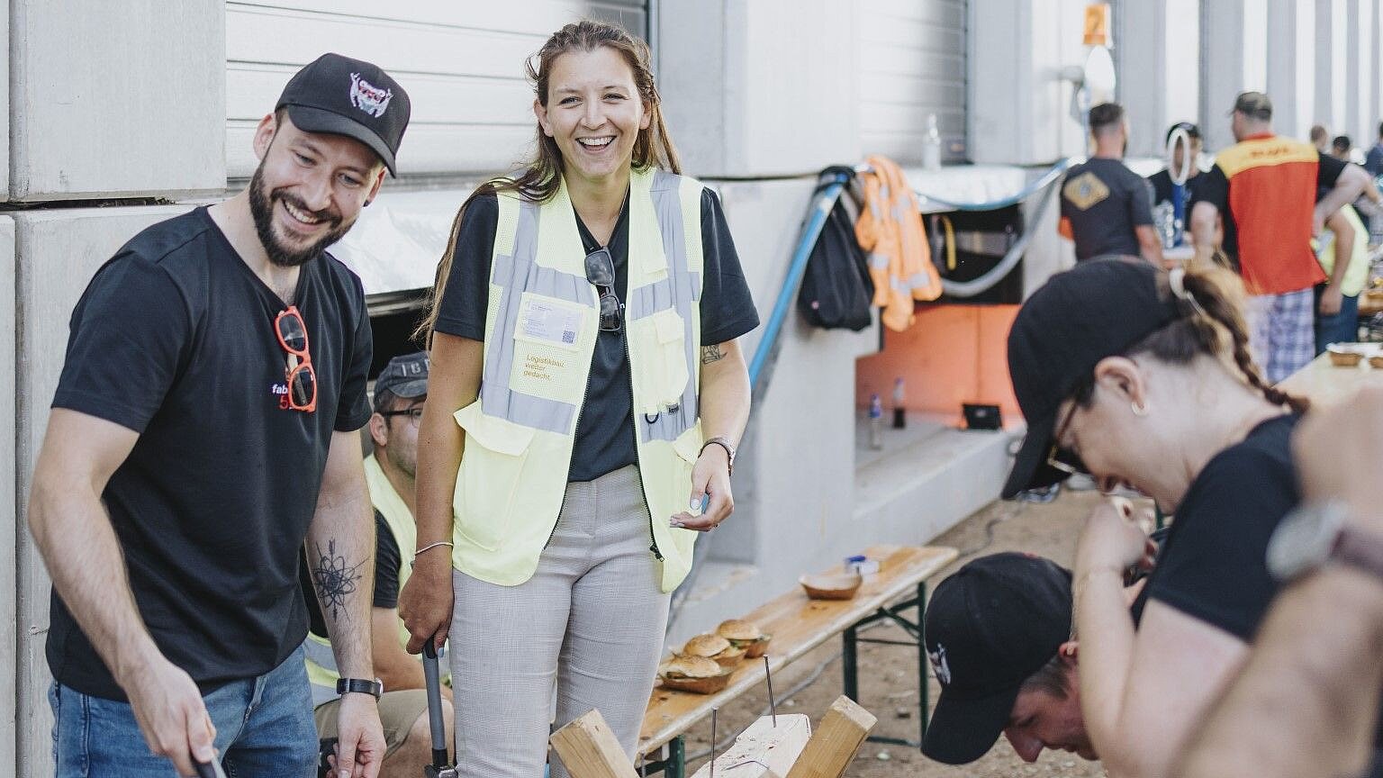 Two smiling people wearing yellow safety vests are working on a wooden structure while others watch in the background.