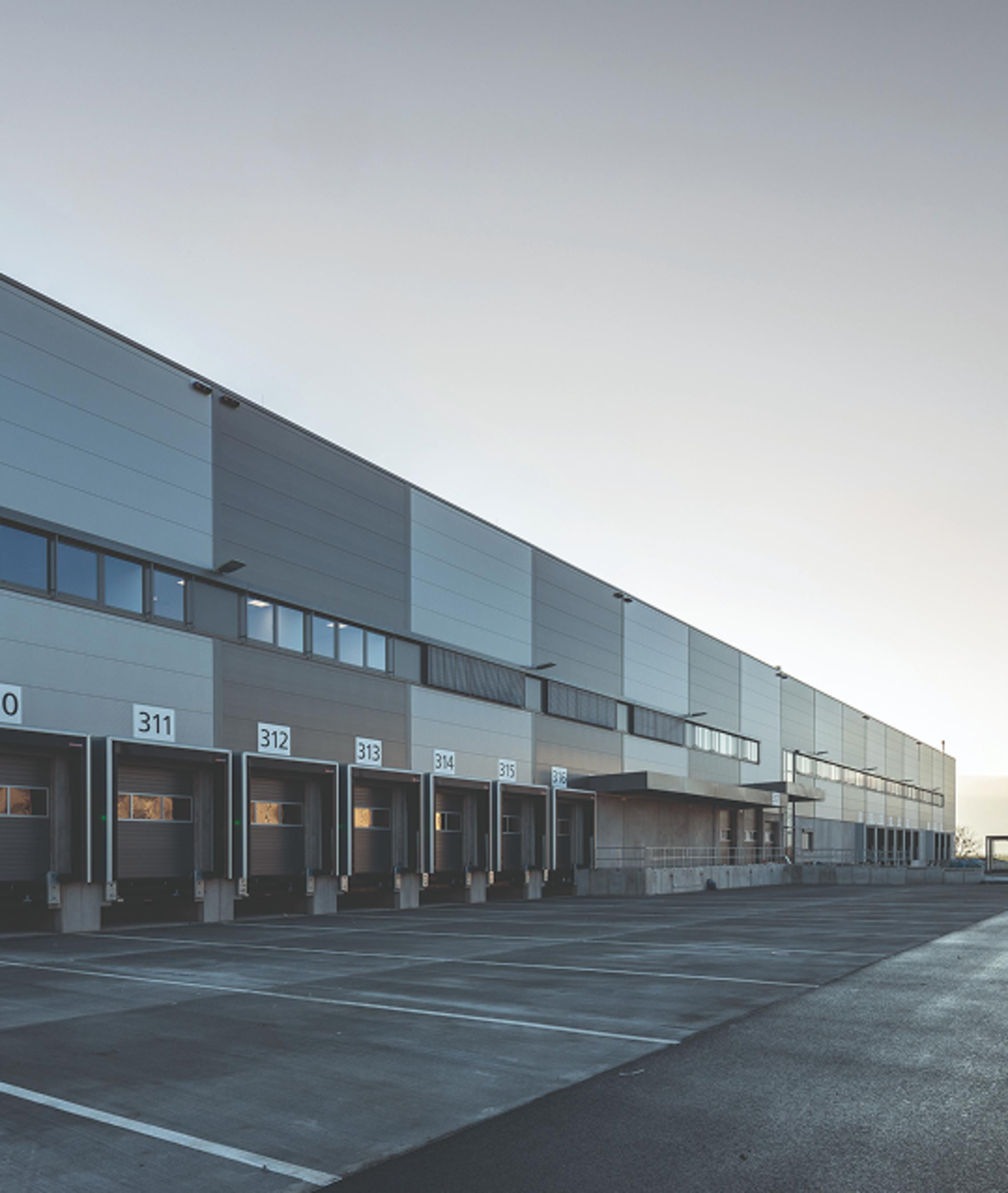 Verbundene Lagerhalle mit mehreren Ladevorrichtungen und grauem Asphaltparkplatz bei Dämmerung.