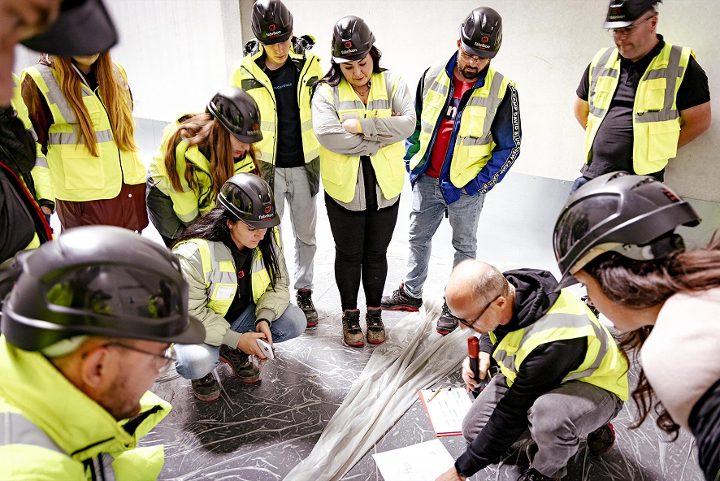 Gruppe von Menschen in Sicherheitswesten und Helmen, die um einen Plan auf dem Boden versammelt sind.