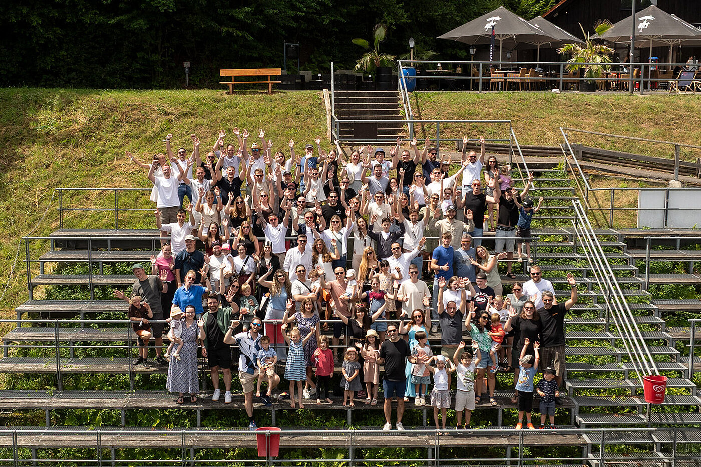 A large group of people stand on bleachers and wave at the camera, surrounded by green nature and an event venue.