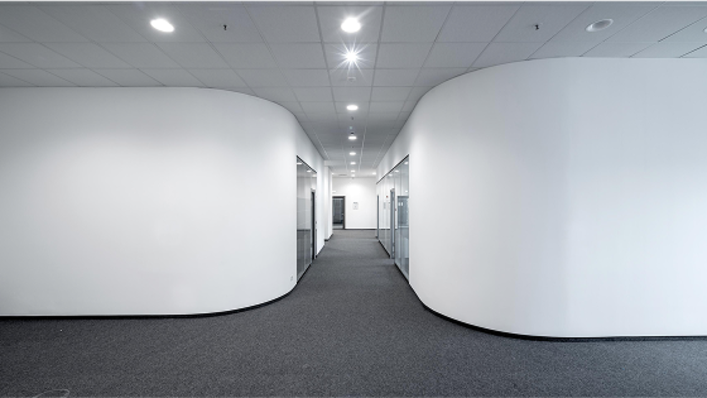 Ein heller Flur mit weißen Wänden, abgerundeten Ecken und Glaswänden an den Seiten, beleuchtet durch Deckenlampen.