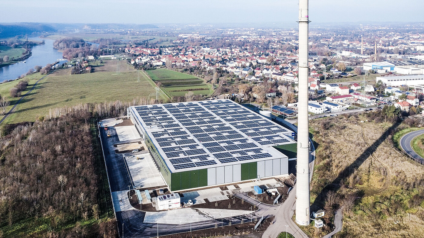 Luftaufnahme eines modernen Gebäudes mit Solarpanelen, umgeben von Landschaft und Wohngebieten.