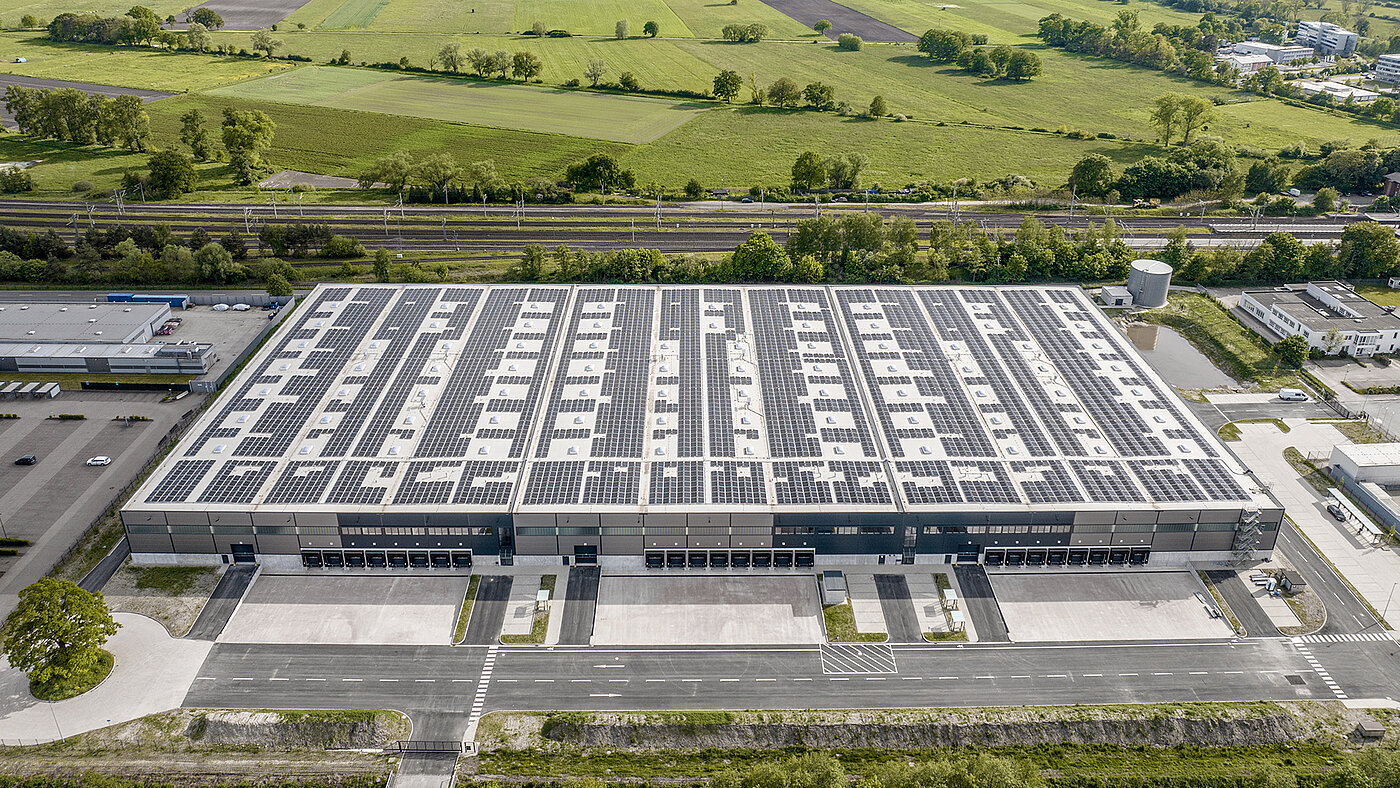 Luftaufnahme eines modernen Industriegebäudes mit Solarpanelen auf dem Dach, umgeben von grünen Feldern und Straßen.