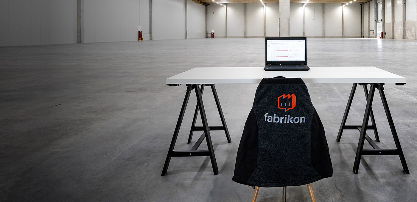 A modern workspace with a white table, a laptop, and a chair with a "fabrikon" logo in an empty room.