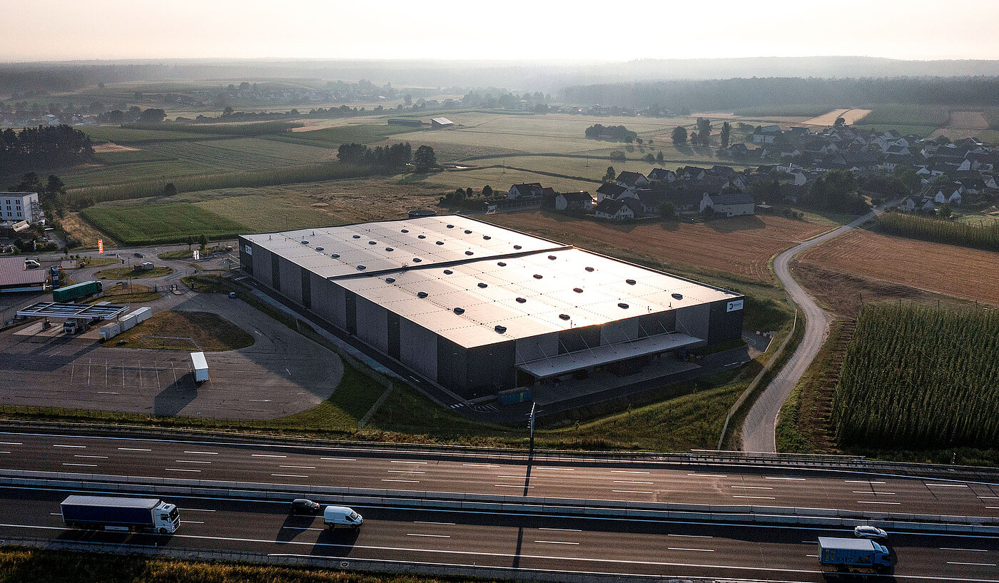 Ein modernes Lagergebäude mit Solarpanelen, umgeben von Feldern und einem sichtbaren Highway im Hintergrund.