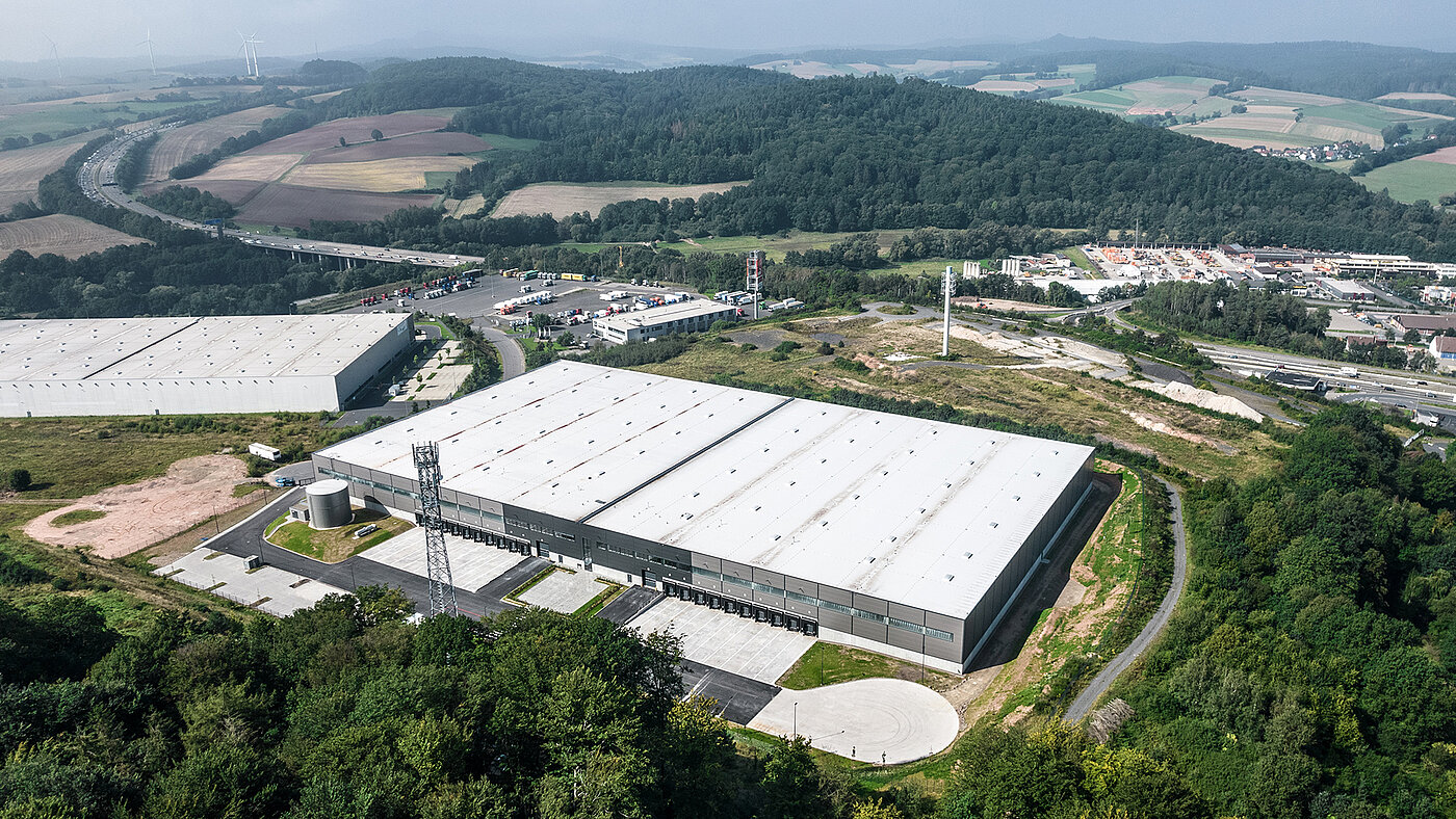 Luftaufnahme eines modernen Logistikzentrums umgeben von Autobahn, Fahrzeugen und ländlicher Landschaft.