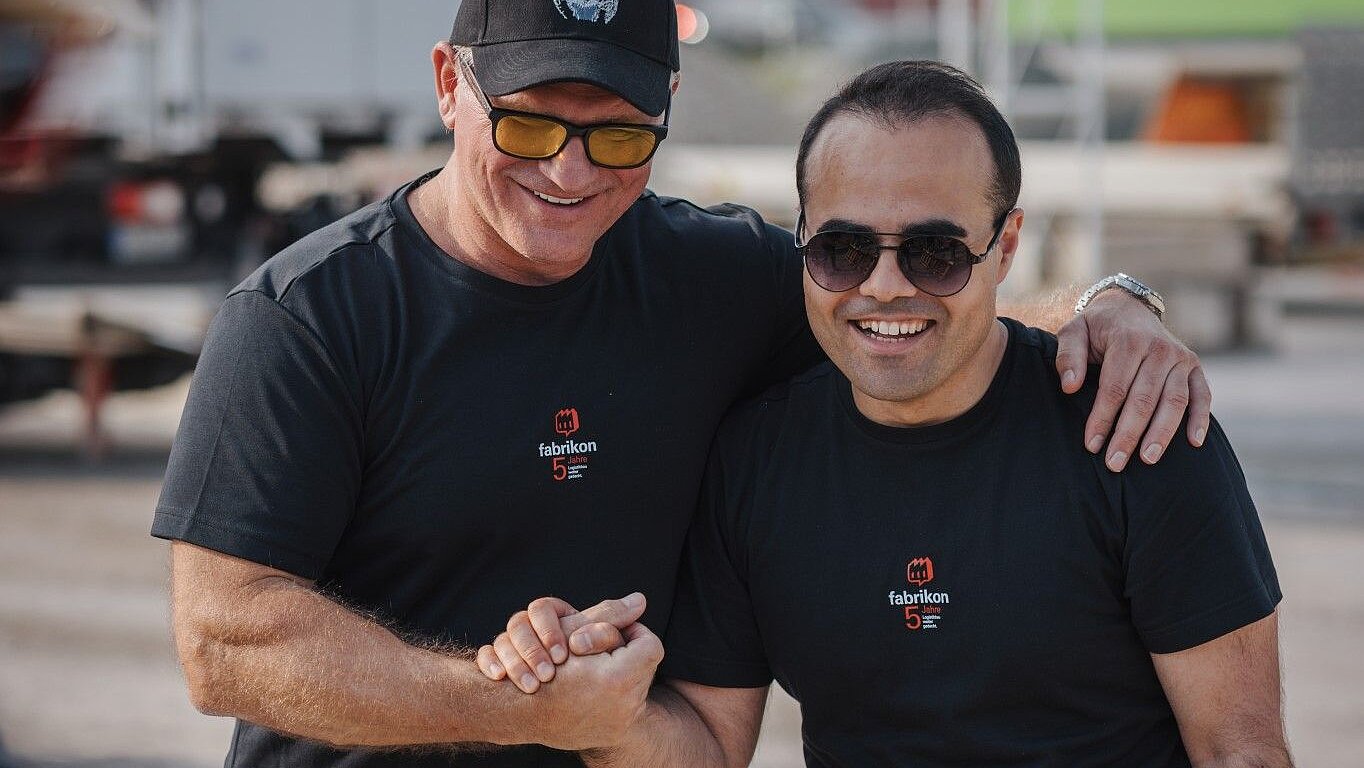 Two men in black T-shirts smile and hold hands in a friendly manner in an outdoor setting.