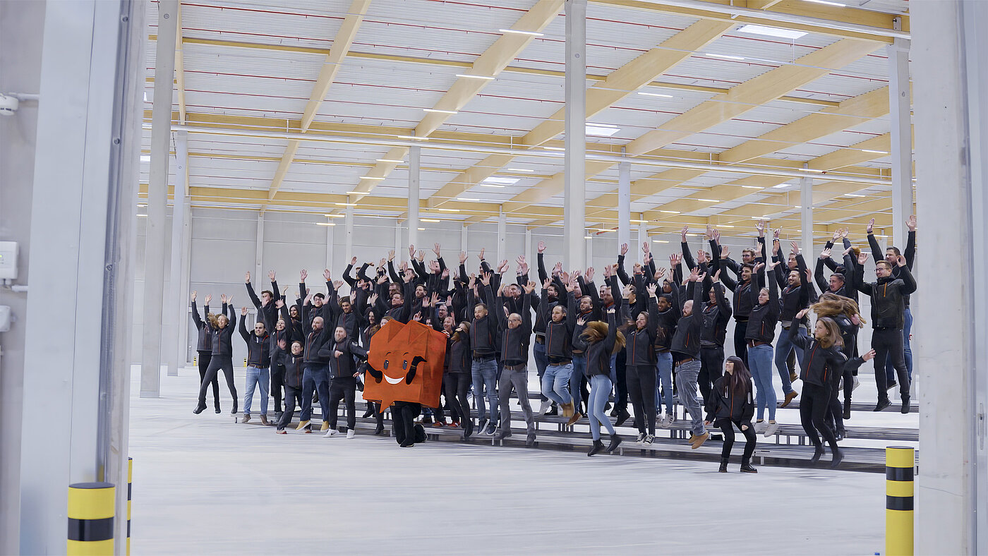 Eine große Gruppe von Menschen in Arbeitskleidung hebt die Hände in die Luft, während sie in einem geräumigen Innenraum stehen.