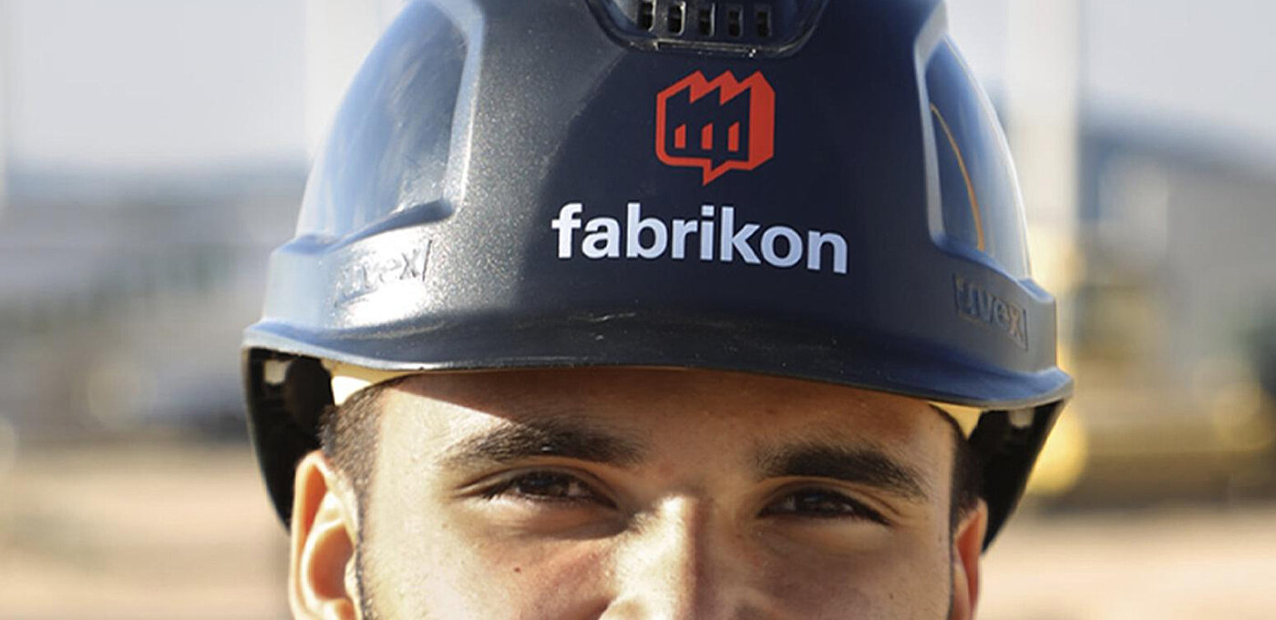 Mann mit Schutzhelm, auf dem das Logo "fabrikon" sichtbar ist, vor unscharfer Baustellenkulisse.