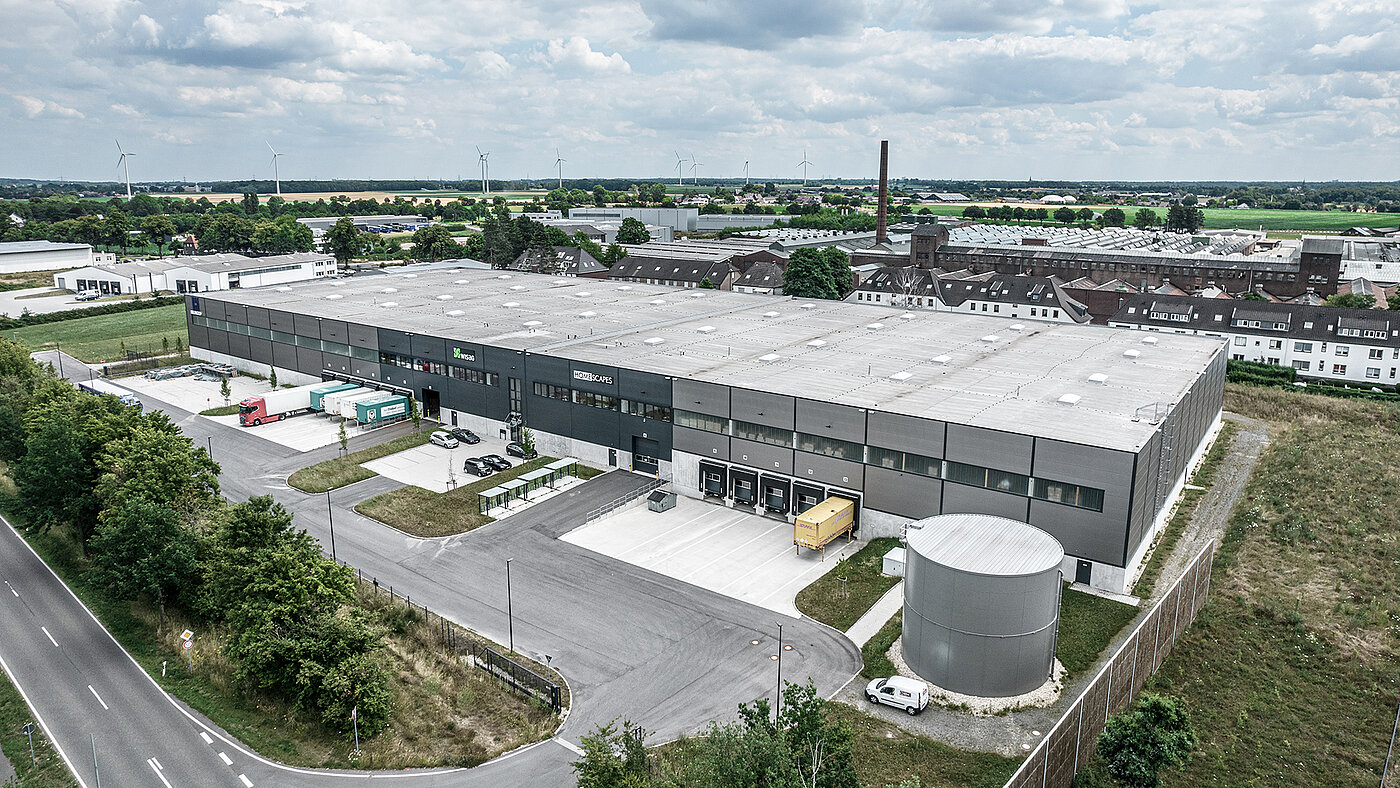 Luftaufnahme eines modernen Lagergebäudes mit Lkw, Pkw und Windkraftanlagen im Hintergrund.