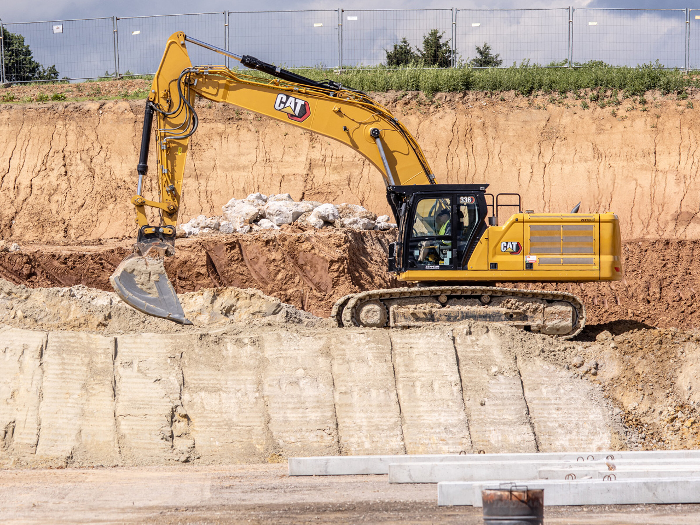 Bagger auf einer Baustelle hebt Erde an, im Hintergrund ein abgetrennter Bereich mit Baumreihe und Bauzäunen.