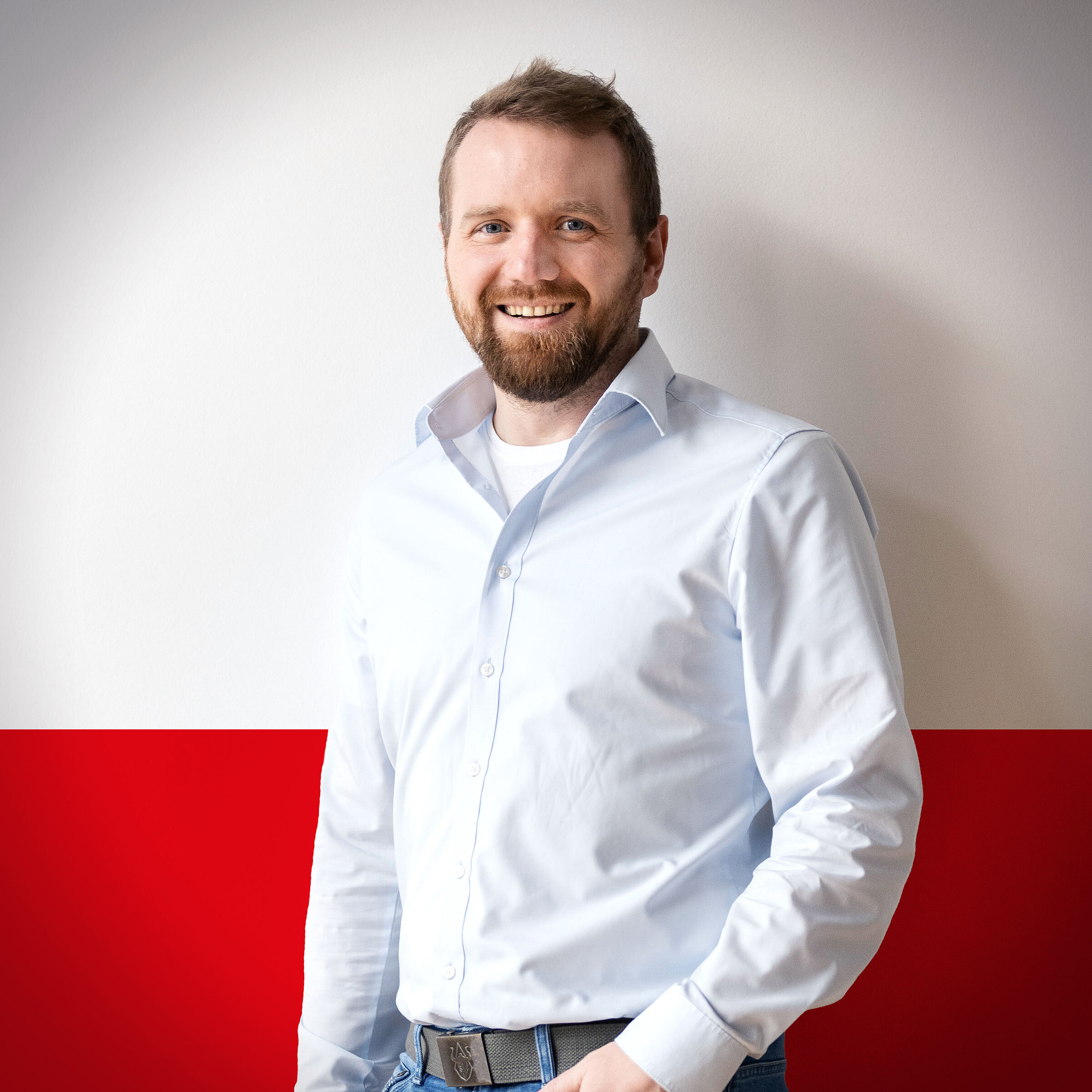 Smiling man in a light blue shirt in front of a red and white background, posing confidently.