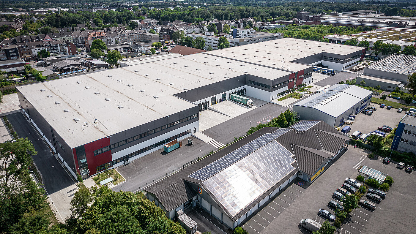 Luftaufnahme eines modernen Logistikzentrums mit Parkplätzen und umliegenden Gebäuden. Solarpanels auf dem Dach.