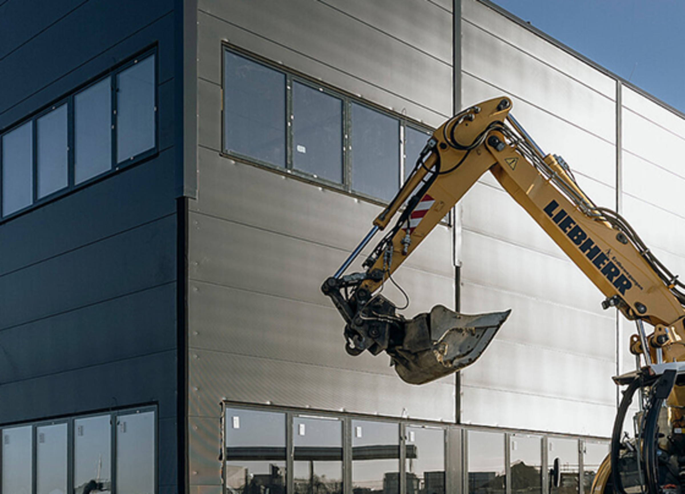 Excavator working on a modern building with large windows and a metal facade.