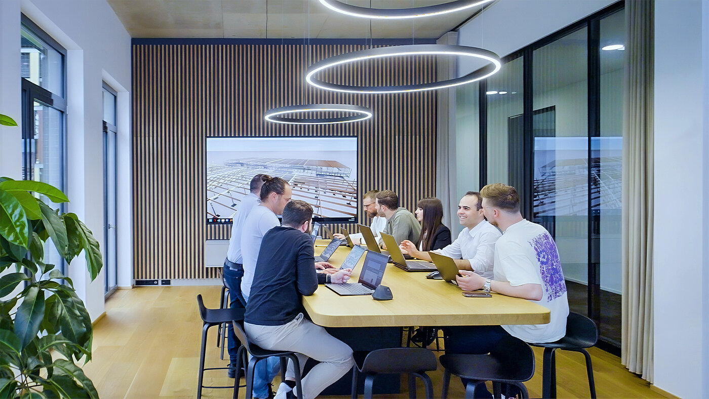 Teammeeting in modernem Raum mit Laptops und Bildschirm, umgeben von Pflanzen und Holzverkleidung.